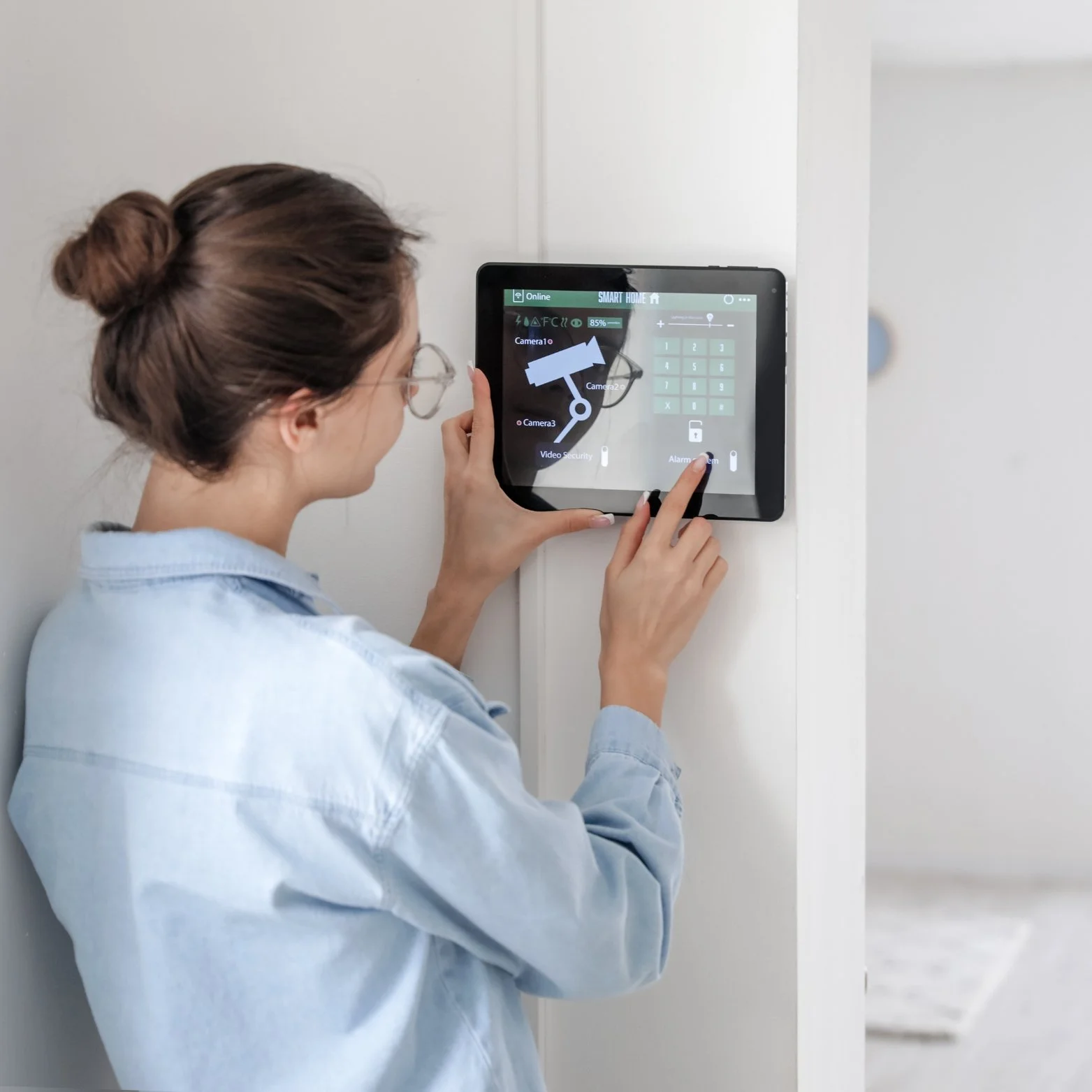 A woman with glasses and a bun hairstyle, wearing a light blue shirt, is operating a home security system on a mounted touchscreen panel.