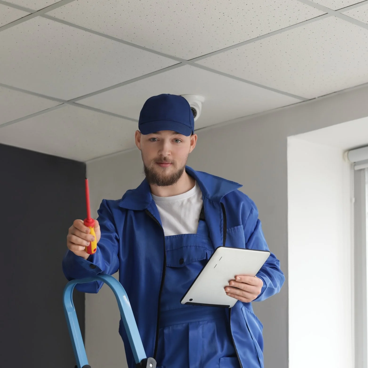 A maintenance worker in a blue uniform and cap holding a screwdriver and a tablet, standing near a ladder indoors.