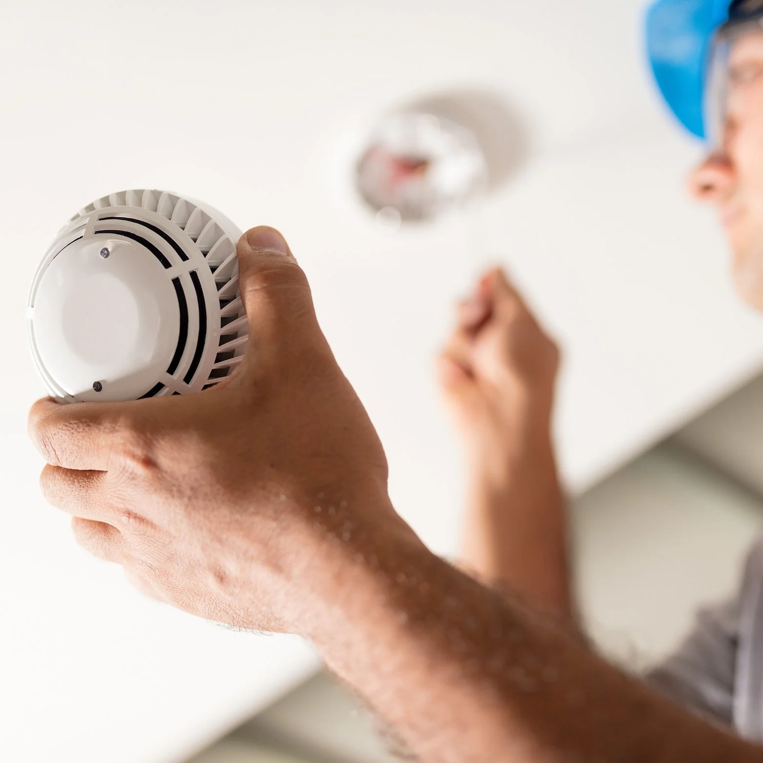 Person installing a smoke detector on a ceiling, wearing safety glasses and a blue hard hat.