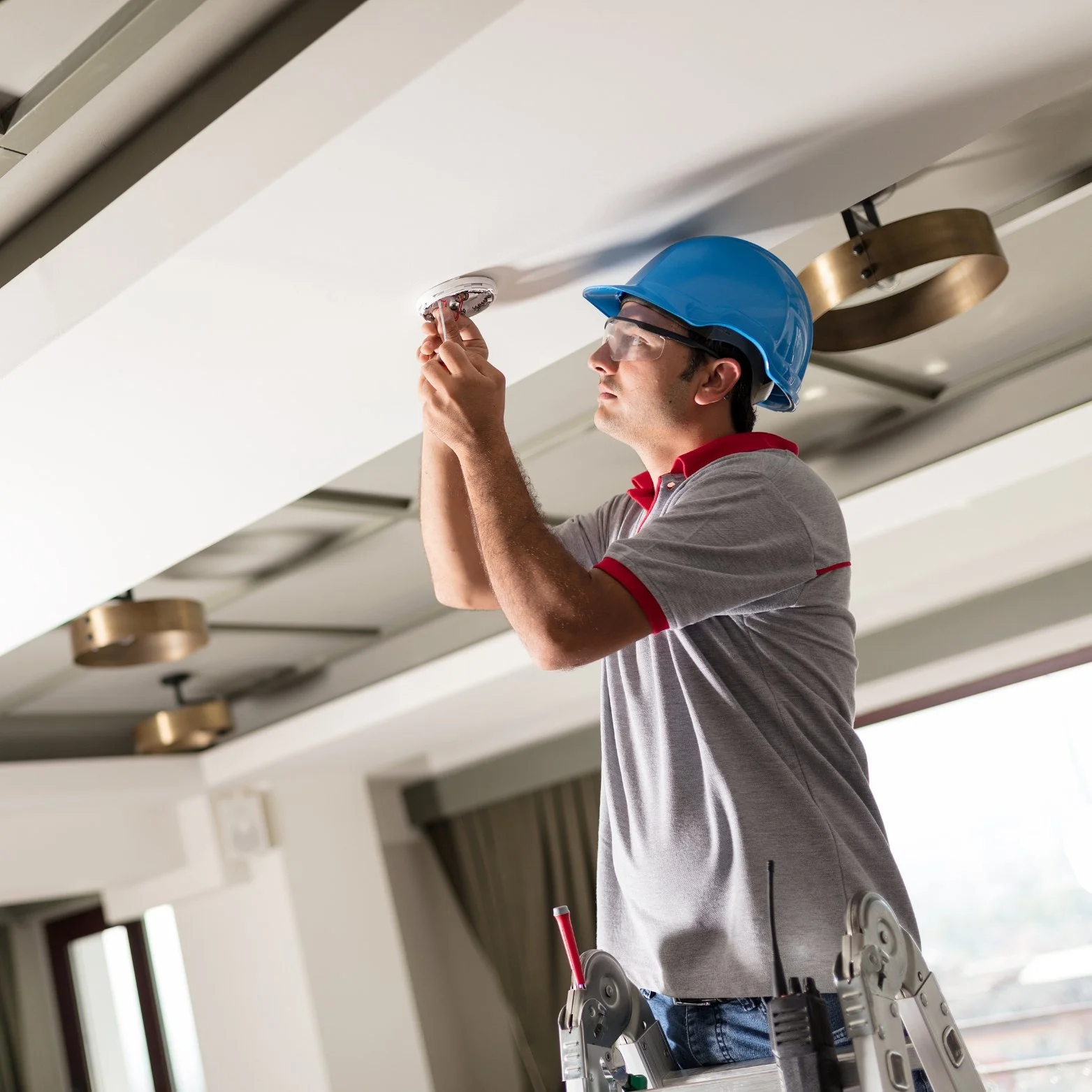 A worker in a blue safety helmet and safety glasses installs a smoke detector on the ceiling of a room.