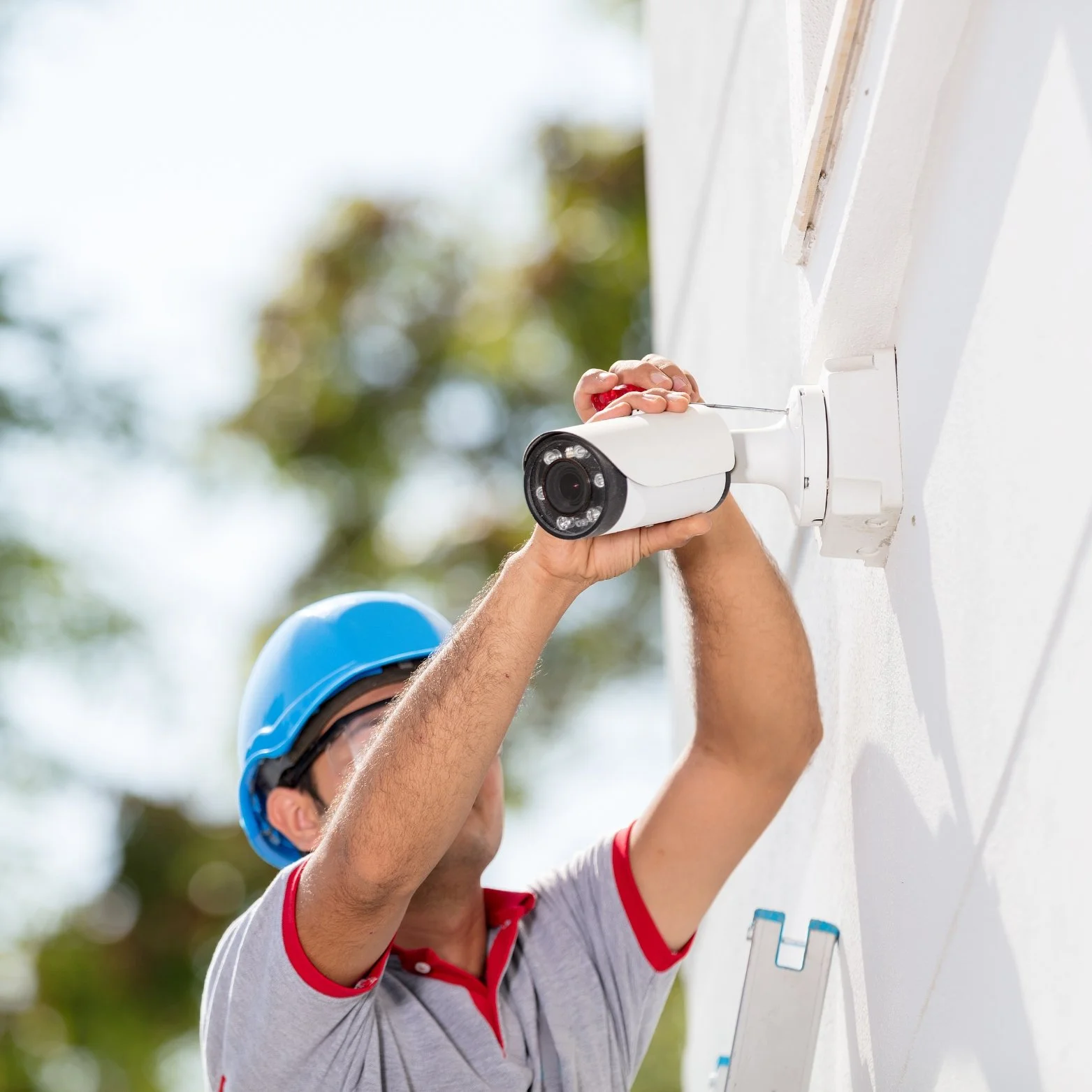 A person wearing a blue hard hat and safety glasses installing a security camera on a white wall outside.