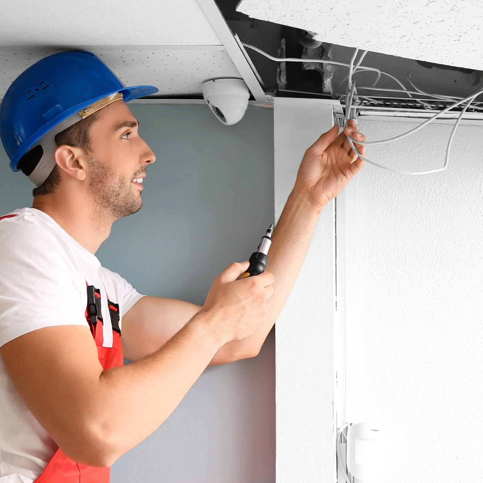 A man wearing a blue hard hat and red suspenders is working on ceiling wiring with a screwdriver.