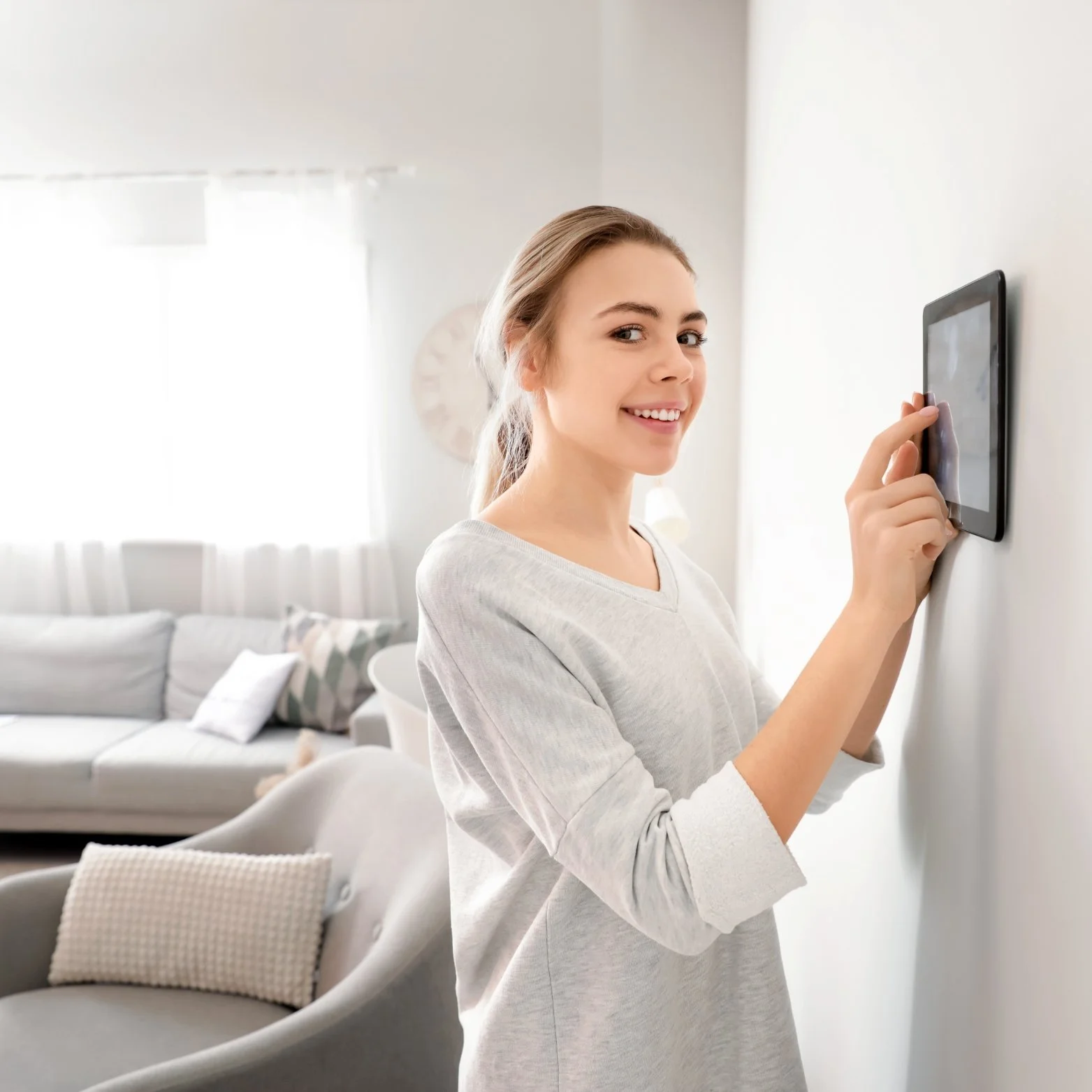 A young woman with light brown hair smiling and pointing at a mounted tablet on a white wall in a bright living room