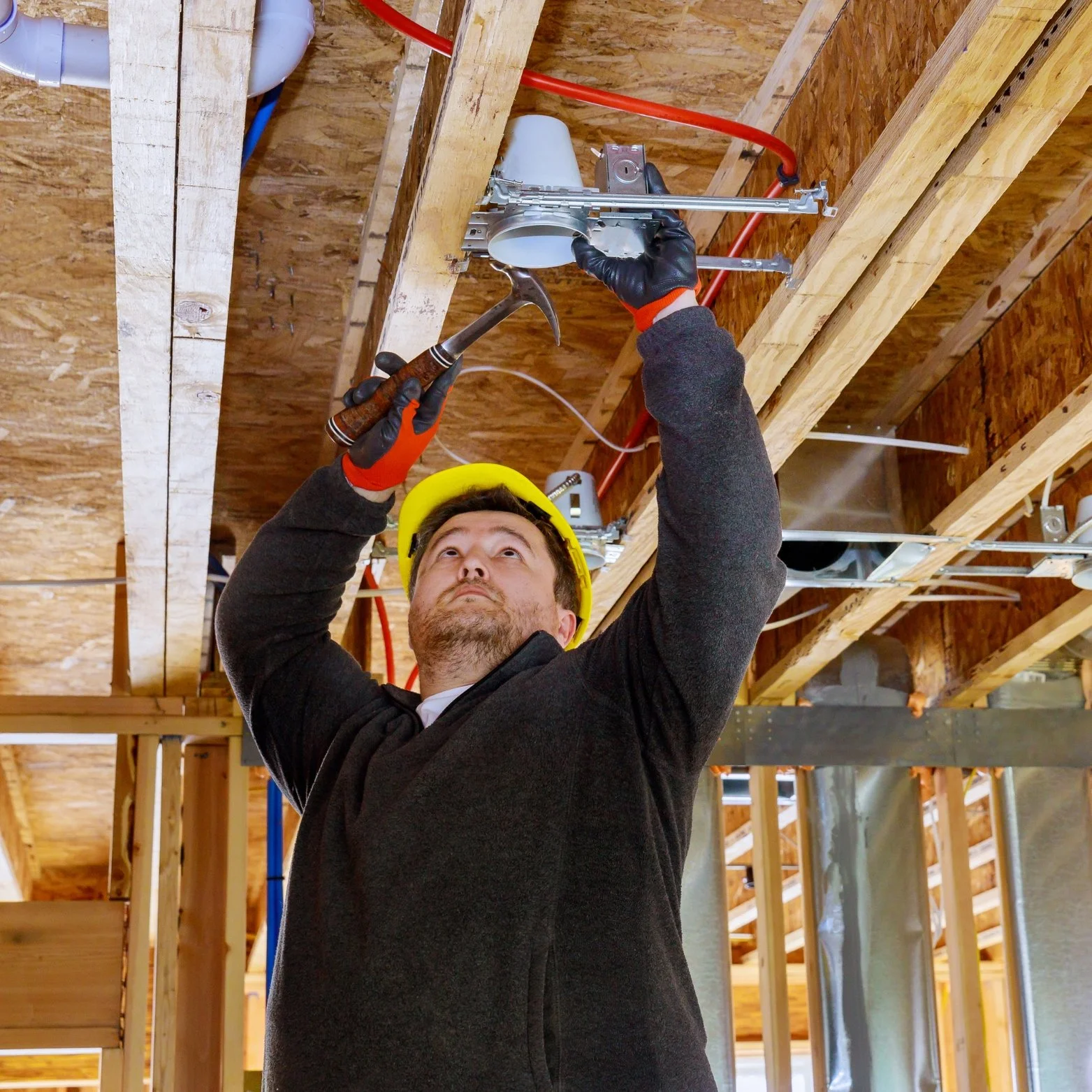 A construction worker wearing a yellow hard hat and gloves using a hammer and a tool to install or adjust a piece of electrical or plumbing conduit on a wooden ceiling framework in a building under construction.