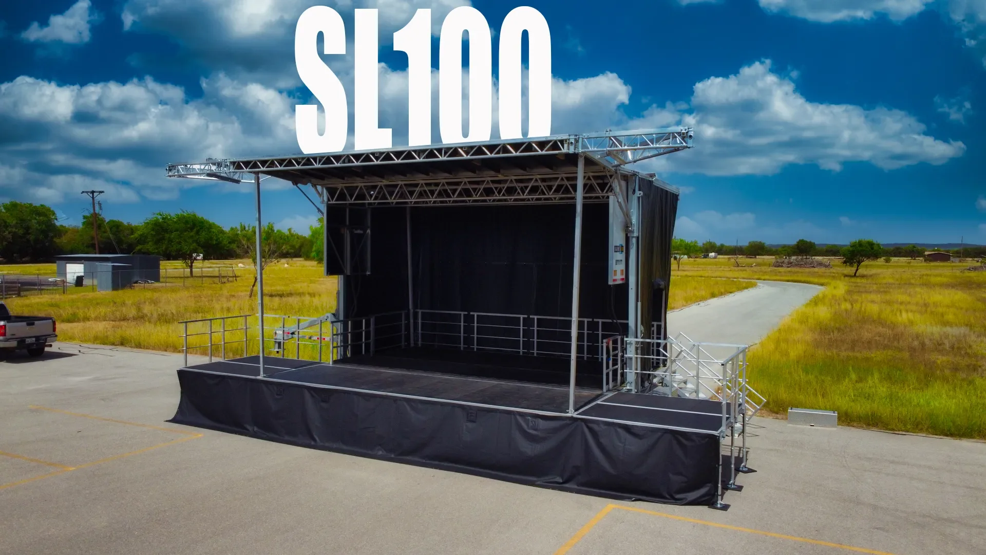 Empty outdoor stage with large $100 sign on top, surrounded by open parking lot and grassy fields under a partly cloudy sky.