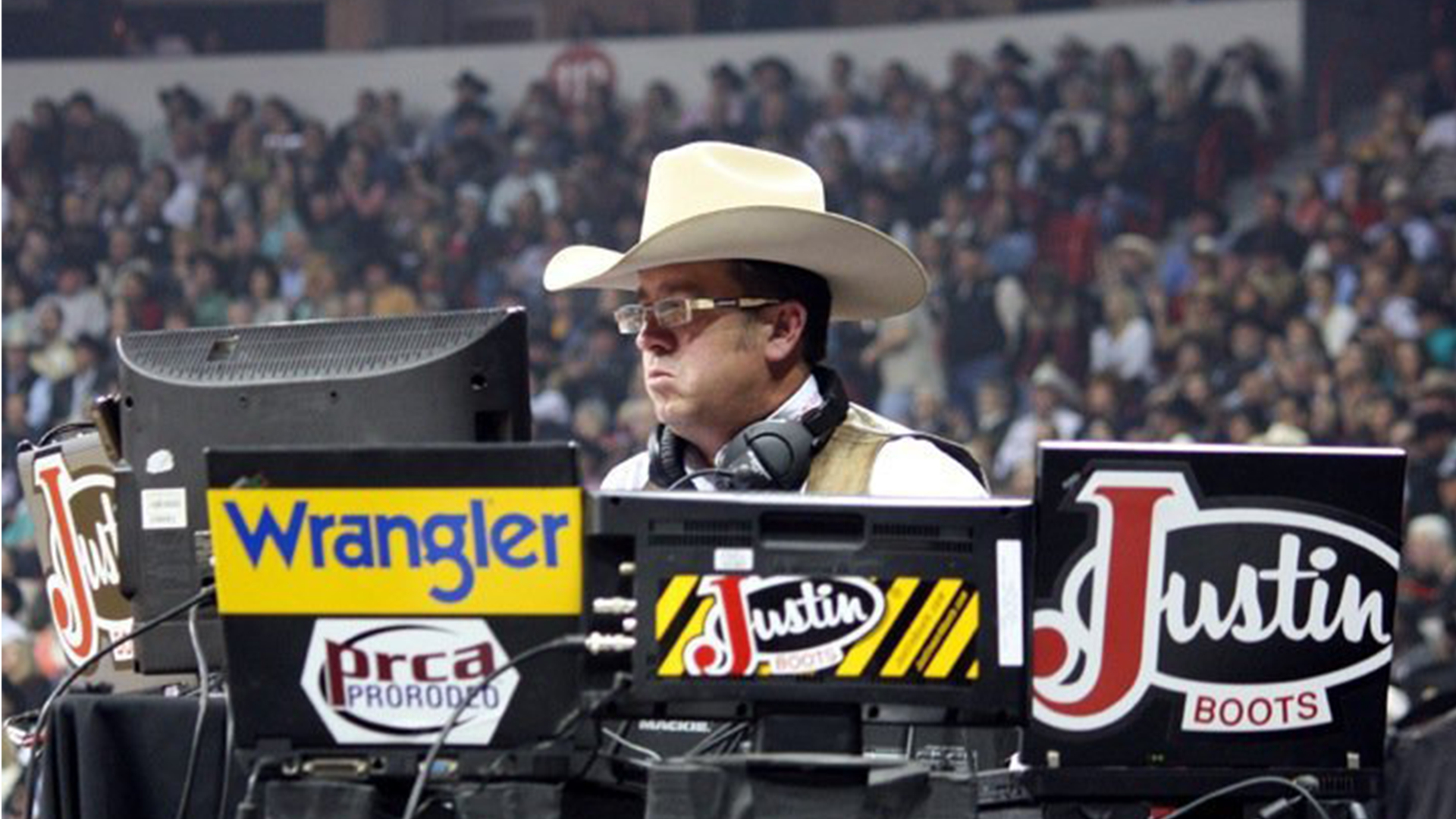 A DJ with a cowboy hat, glasses, and headphones performing at an outdoor event with a crowd in the background.