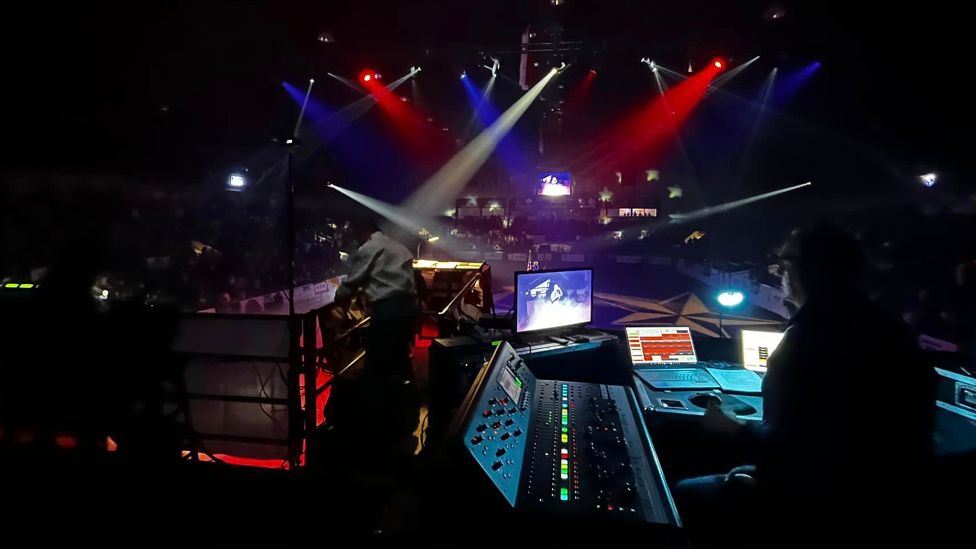 View of a concert or event venue with stage lighting in red, blue, and white, and a sound mixing board and technicians operating sound equipment in the foreground.