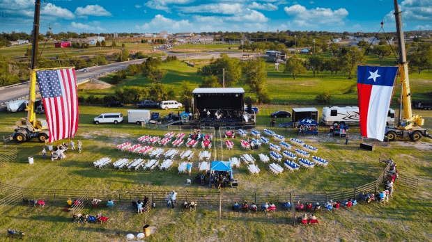 Outdoor event with a stage between two large American and Texas flags, surrounded by folding chairs arranged in rows for attendees, in a grassy field with a small fence and a few people walking around.