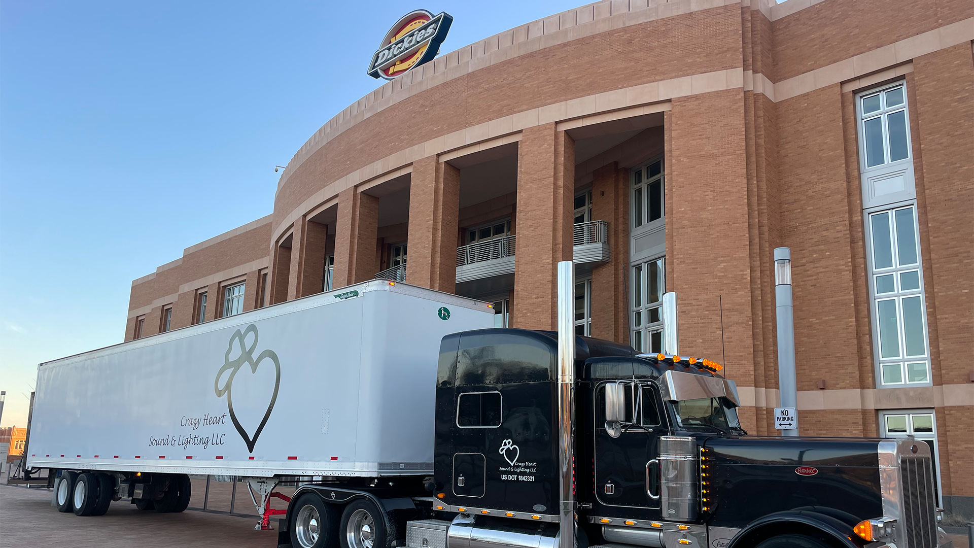 A black semi-truck with a white trailer parked in front of a brick building with large arch windows. The trailer has a logo of a heart with a bow on top and the text 'Crazy Heart Sound & Lighting LLC'. The building has a sign on the roof that says "D