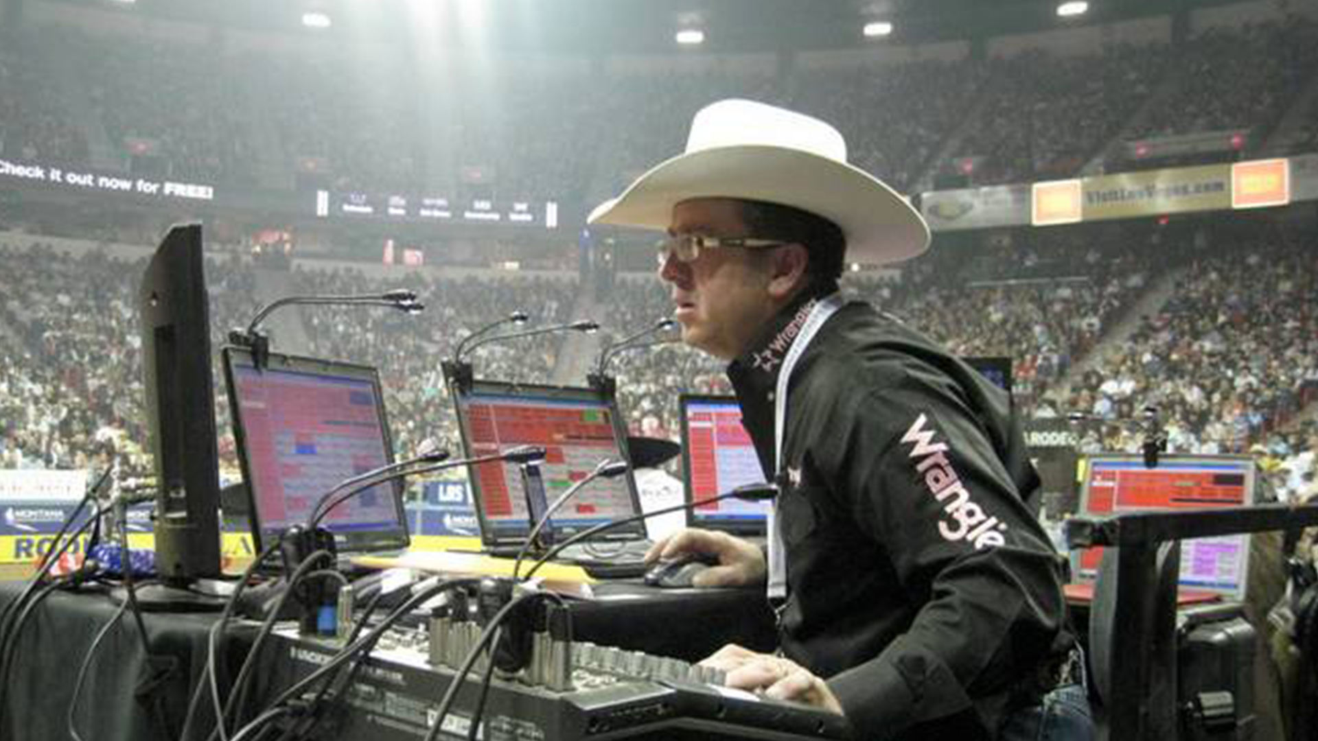 A man in a cowboy hat and glasses operating a sound mixing console at a rodeo event, with a large crowd in the background.
