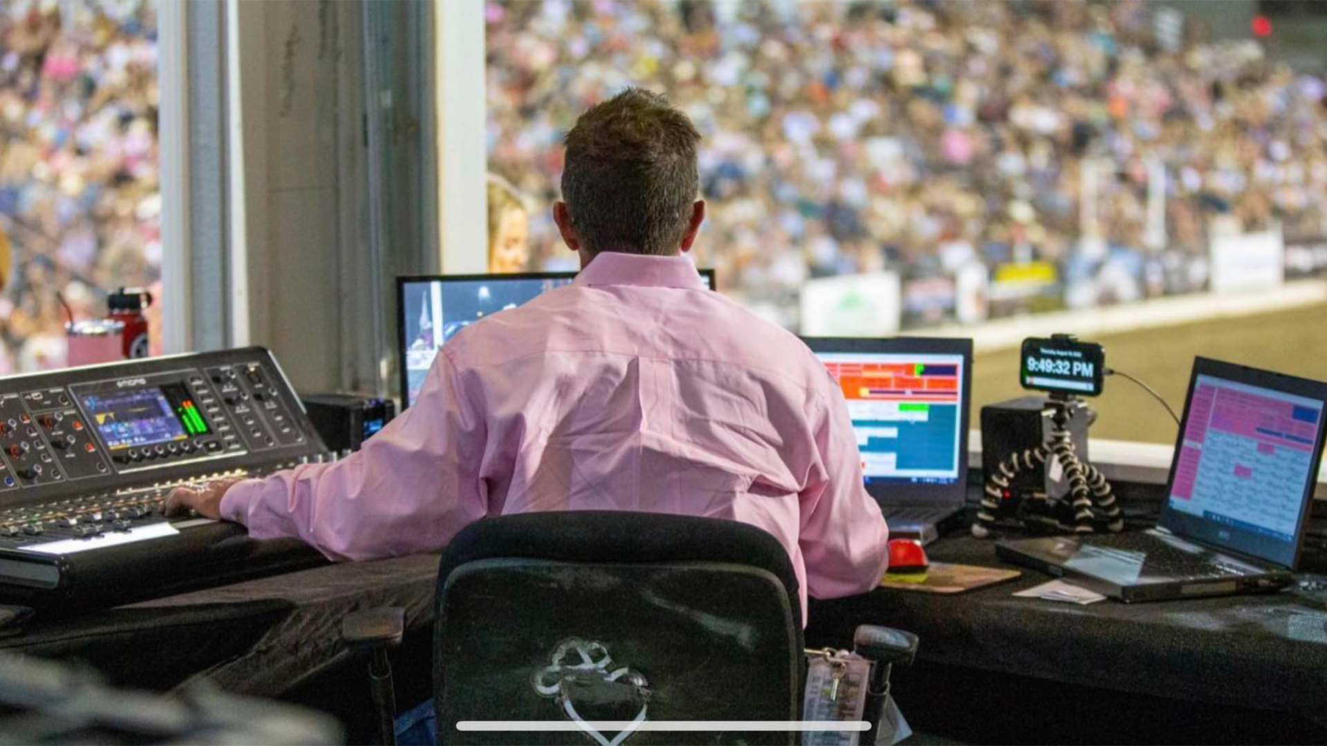 A person in a pink shirt sitting at a control desk with multiple computer monitors and audio equipment, overlooking a large crowd at a sports stadium.