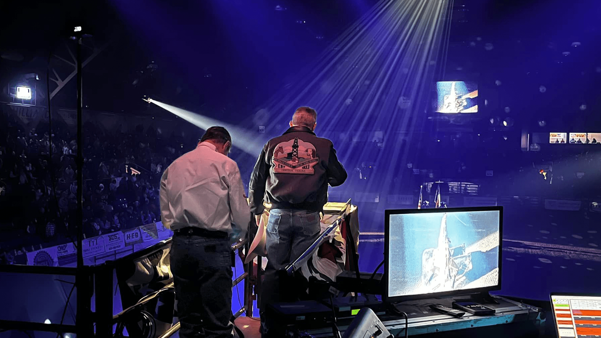 Two men working in a booth at a rodeo or indoor arena, with a large crowd in the background, bright stage lights, and a large screen showing a rodeo event.