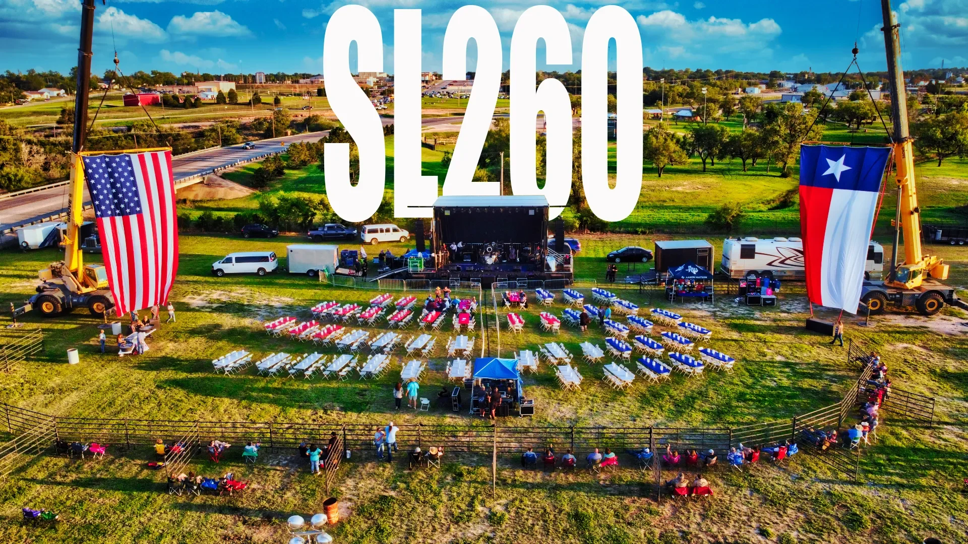 Outdoor concert setup with a stage, seating area with pink, white, and blue chairs, large American and Texas flags on crane towers, and people gathering around on a grassy field under a partly cloudy sky.
