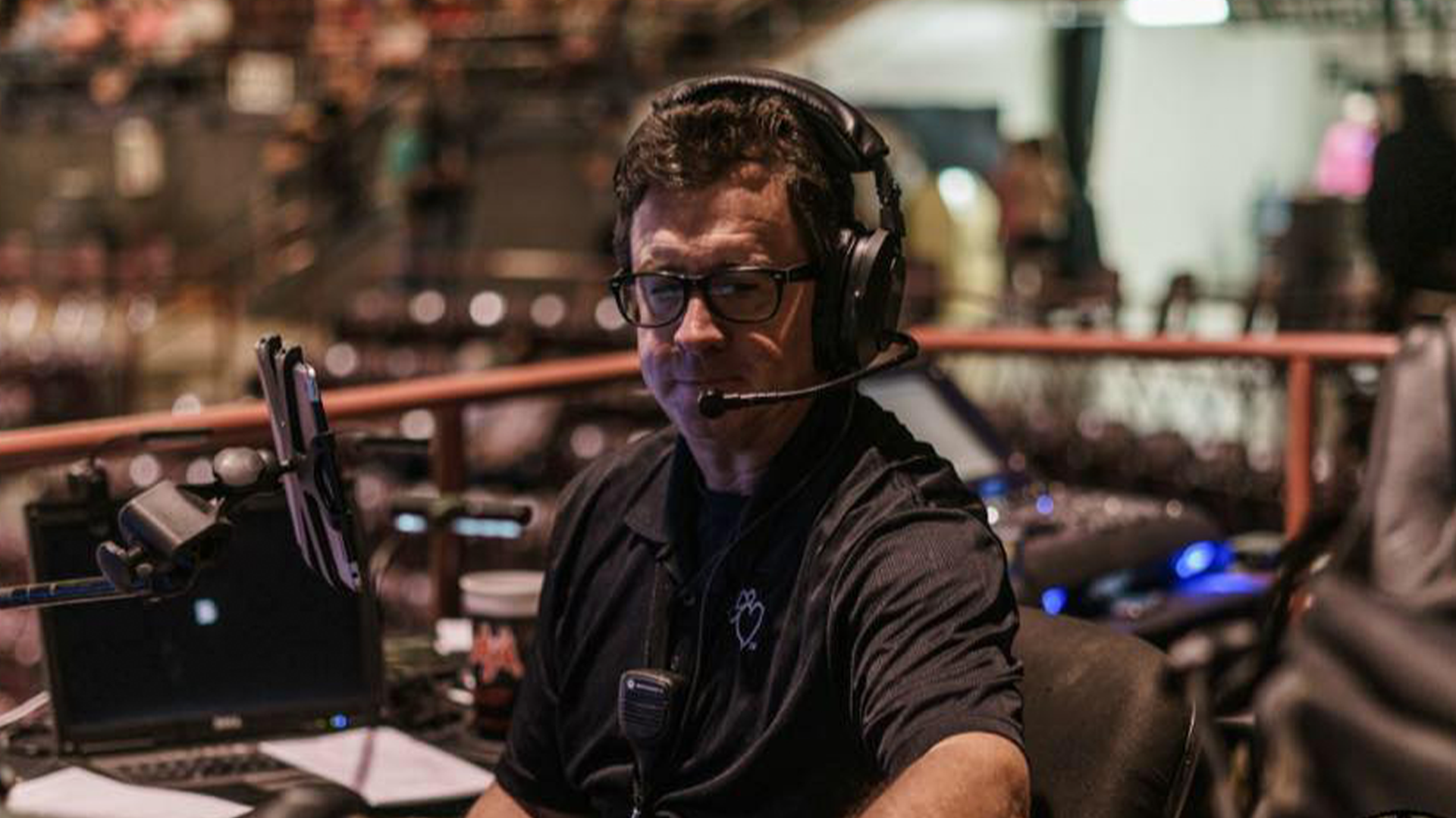 A man wearing glasses and a headset with a microphone, sitting at a DJ or sound mixing station with equipment and a computer in a venue with blurred background.