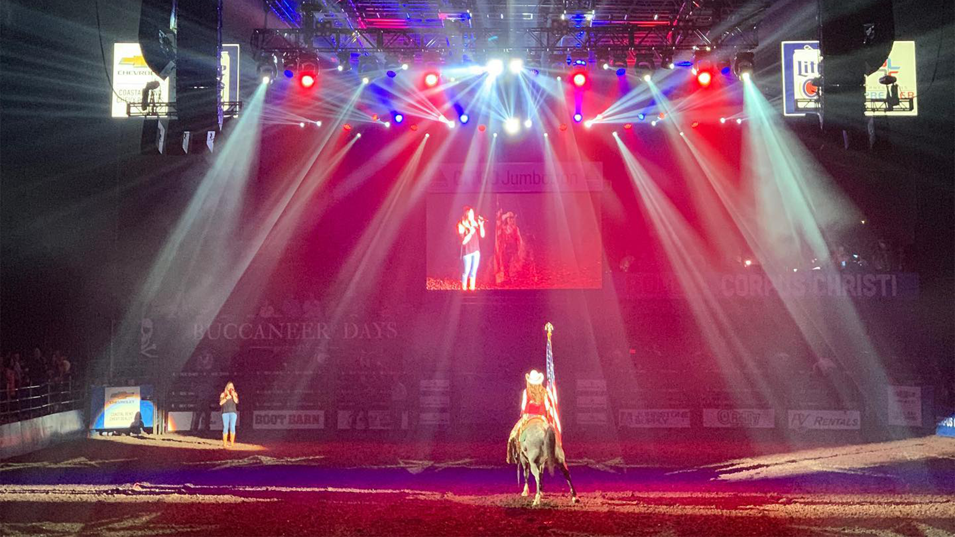 A person riding a horse in a rodeo arena, illuminated by colorful stage lights with a large screen above showing a woman singing.