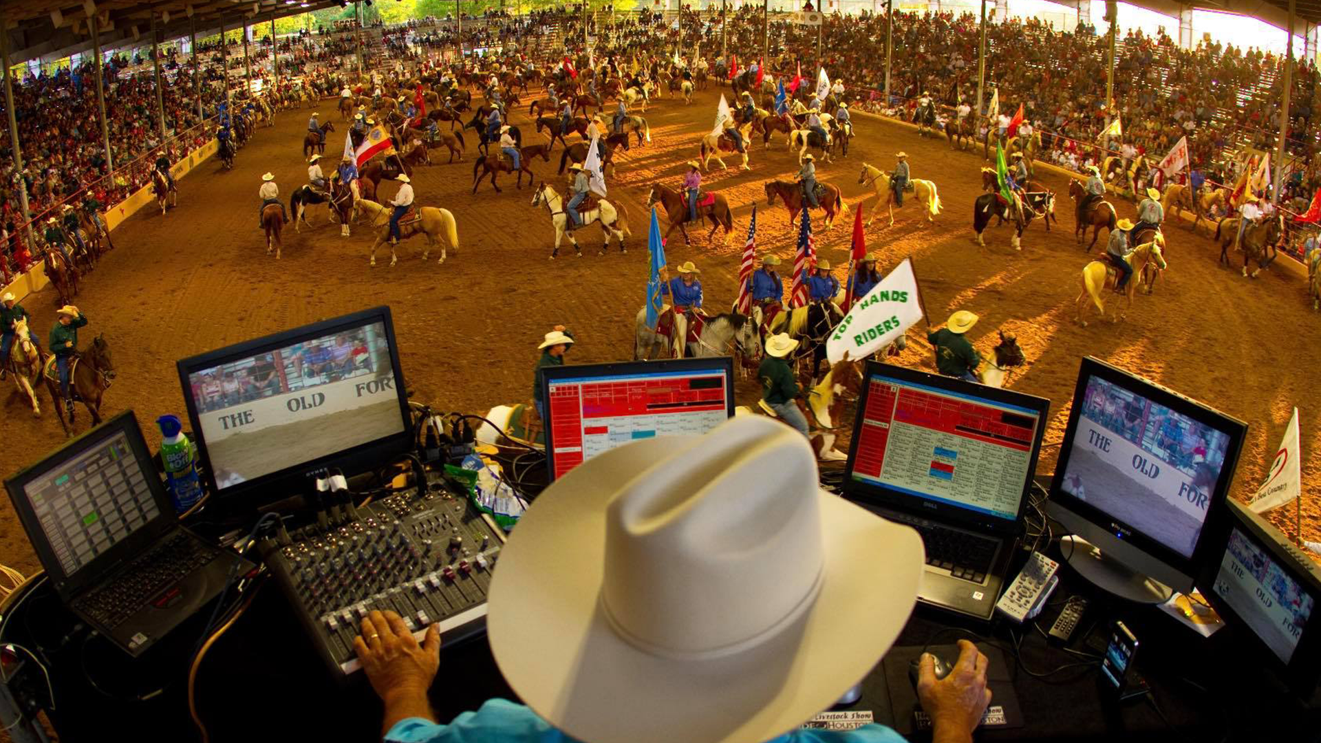 Overview of a rodeo arena with cowboys on horseback, many flags, and a large crowd, viewed from the announcer's booth with a person operating sound and video equipment.
