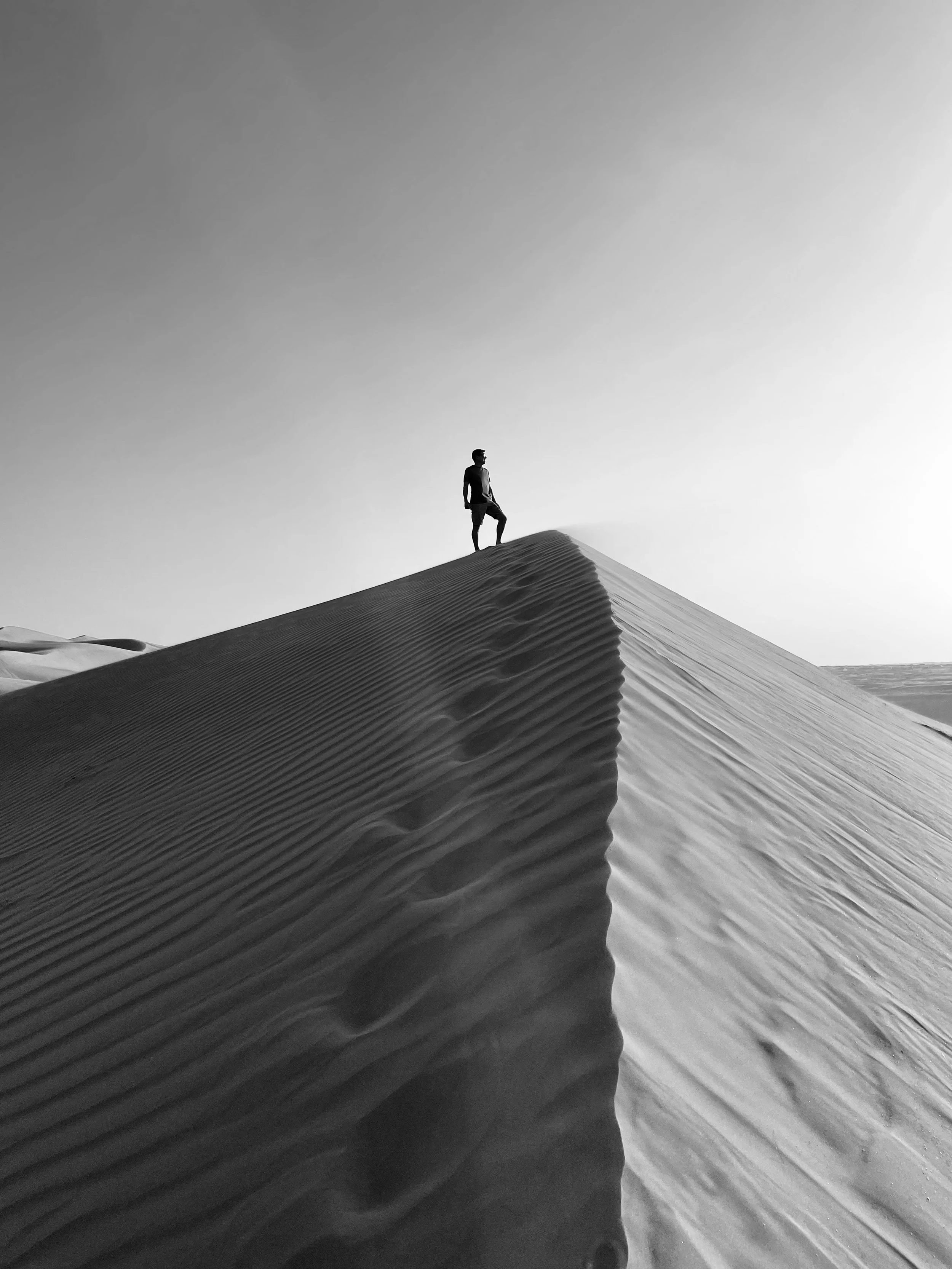 A person standing on the crest of a sand dune in a desert, with footprints trailing behind, captured in black and white.