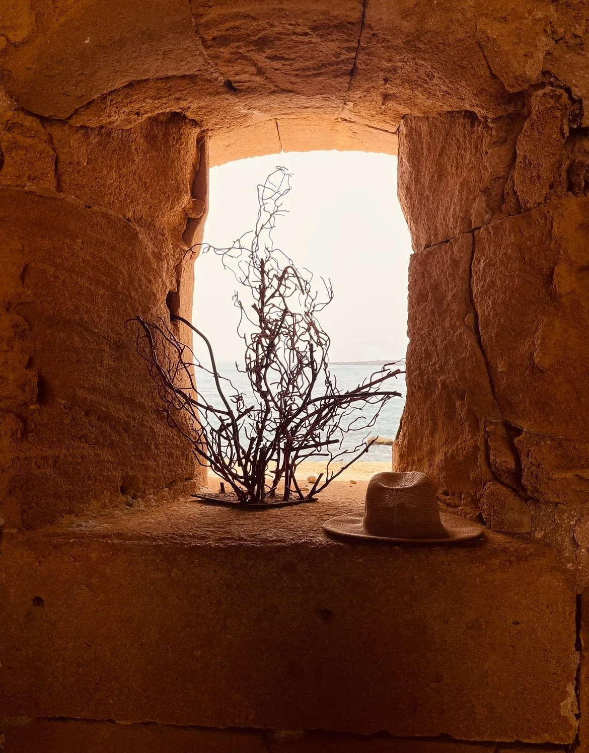 View of a beach seen through a stone window, with a dry branch and a sunhat resting on the stone ledge inside the window.