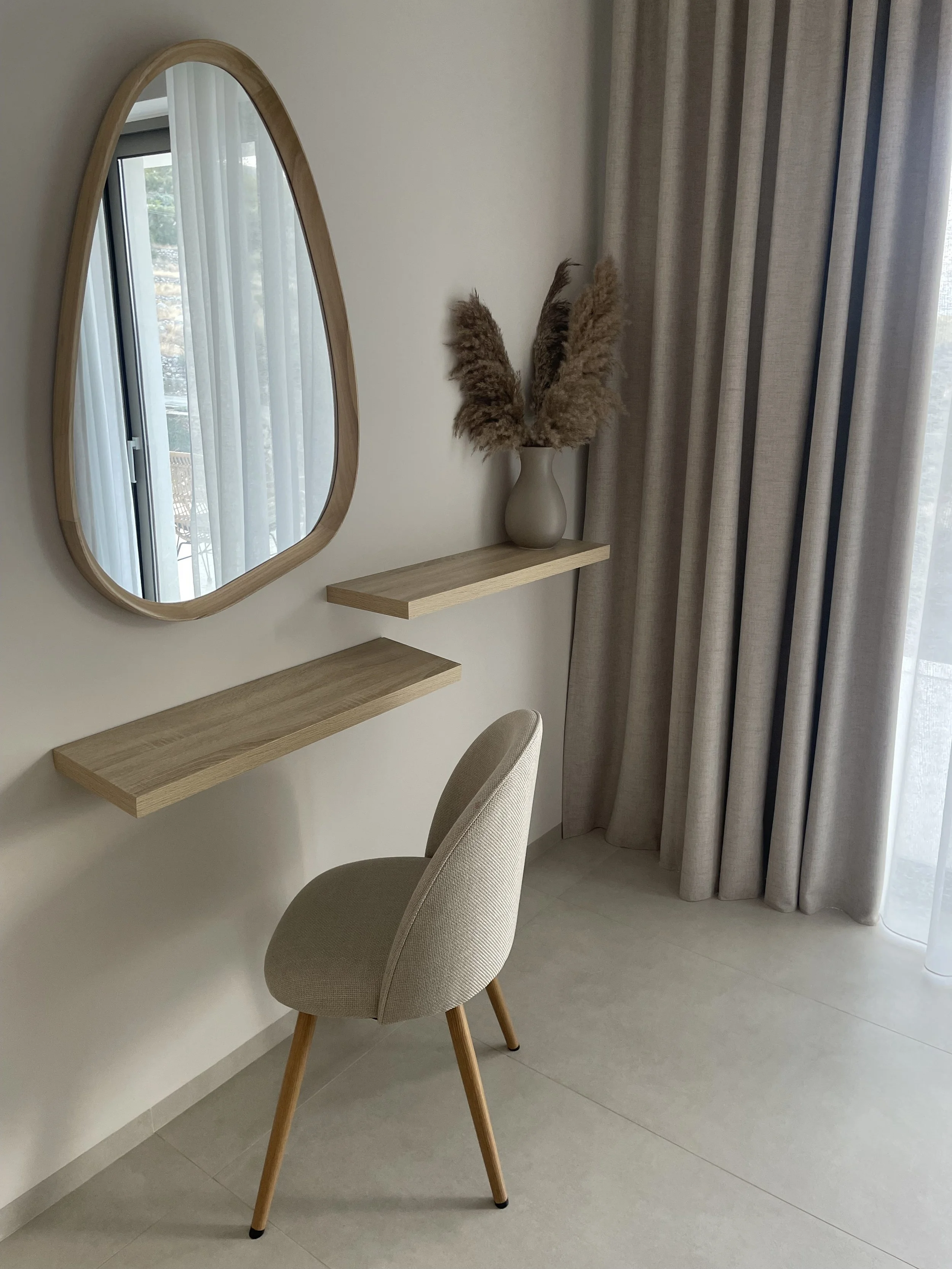 A minimalist vanity corner with an oval mirror, a beige chair with wooden legs, two floating wooden shelves, and a vase with pampas grass near a window with beige curtains.