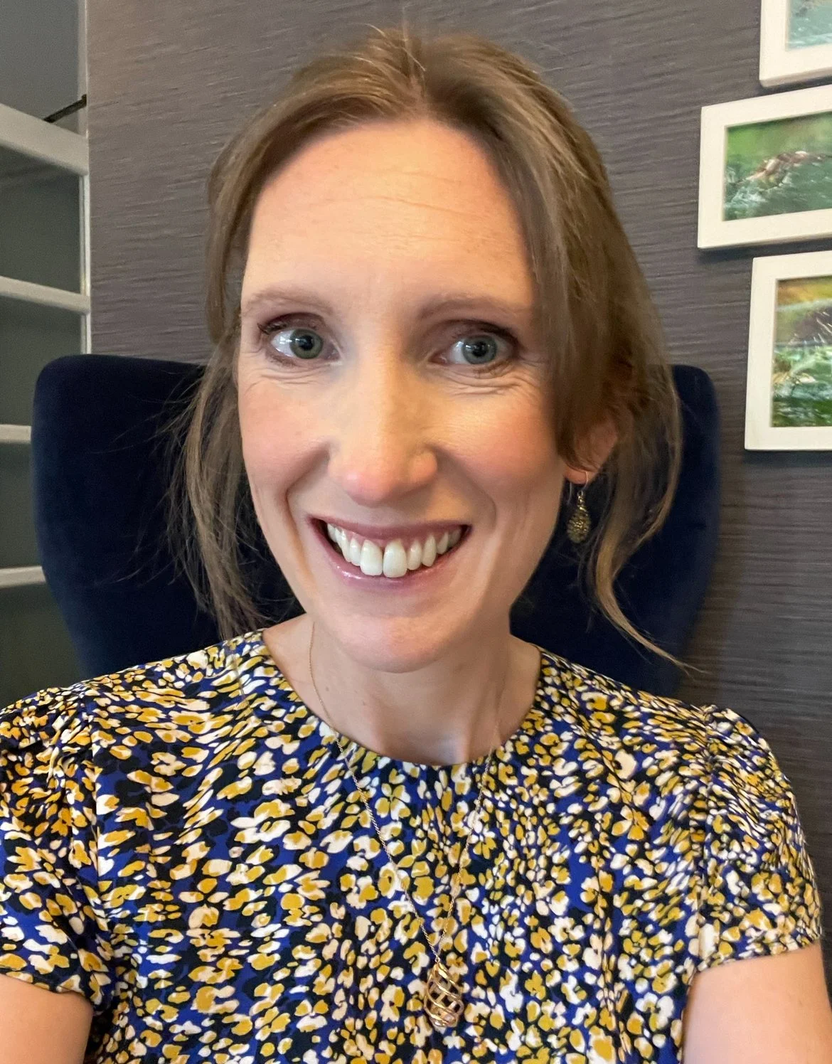 A woman with short brown hair and blue eyes, smiling and wearing a blue, yellow, and white patterned blouse, sitting in front of a dark grey wall with framed pictures, in an indoor setting.