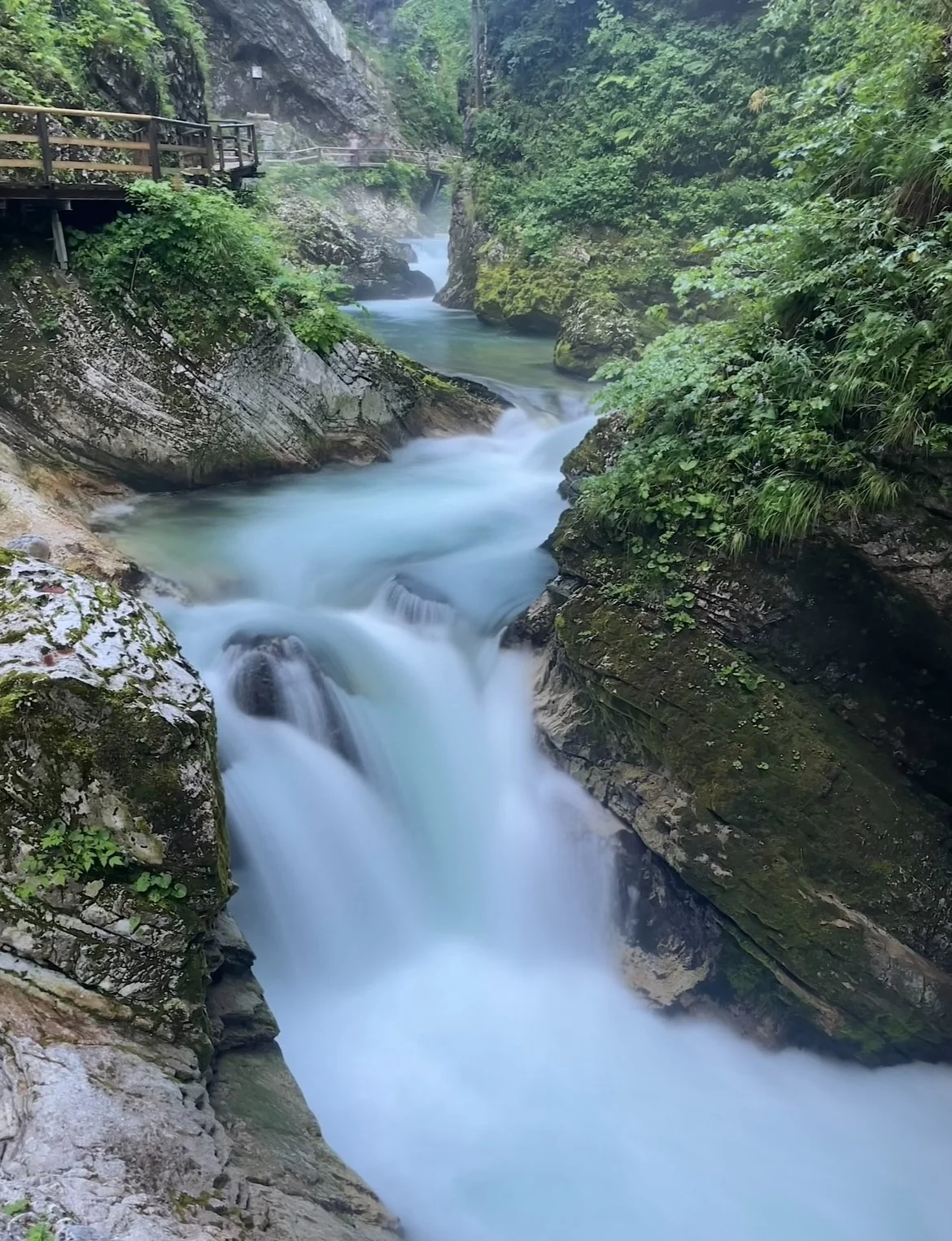 A scenic view of a narrow river cascading over rocks in a forested canyon with a wooden walkway on the left side.