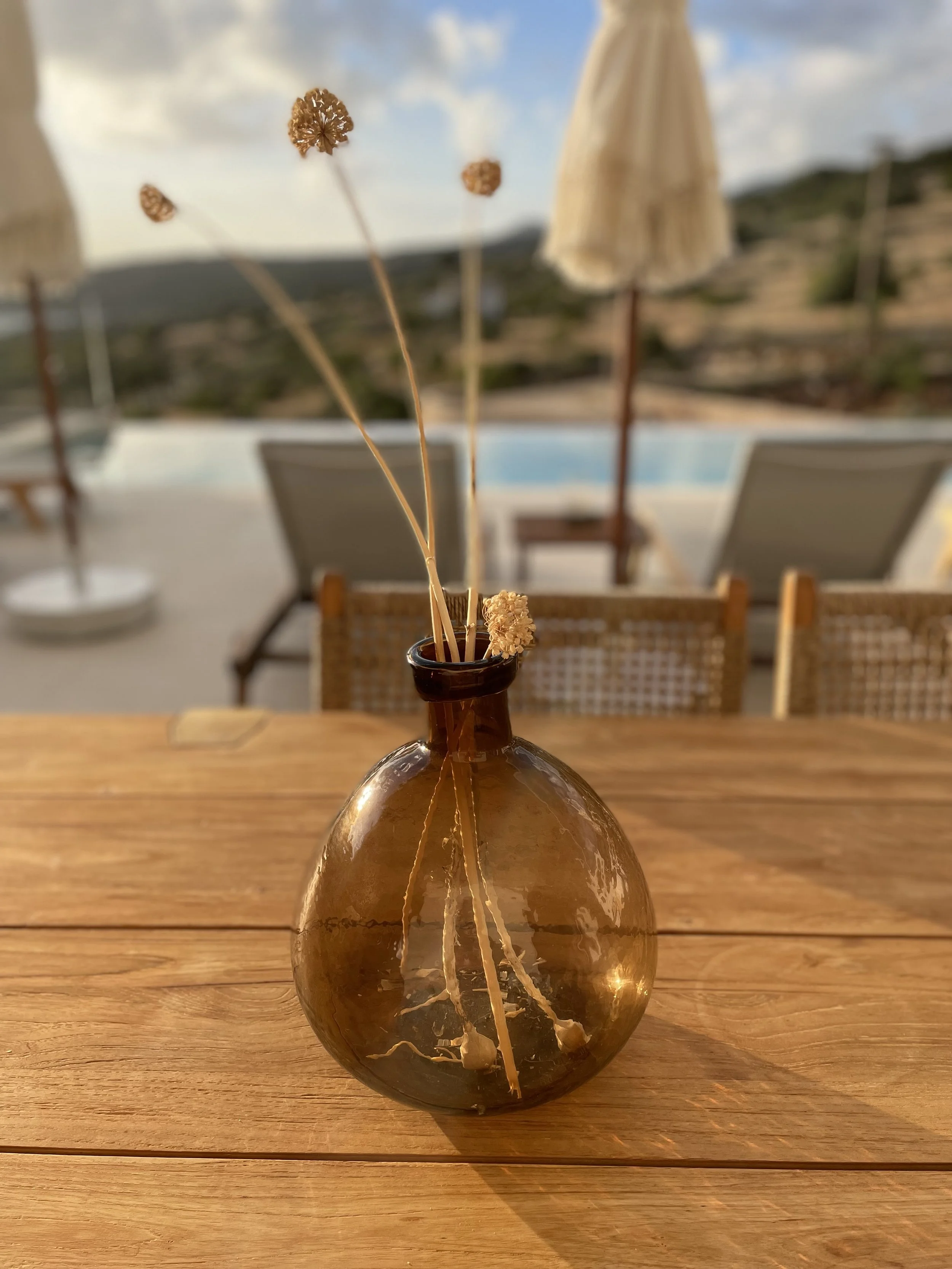 A brown glass vase with dried flowers on a wooden table outdoors with a pool, lounge chairs, umbrellas, and a mountainous landscape in the background.