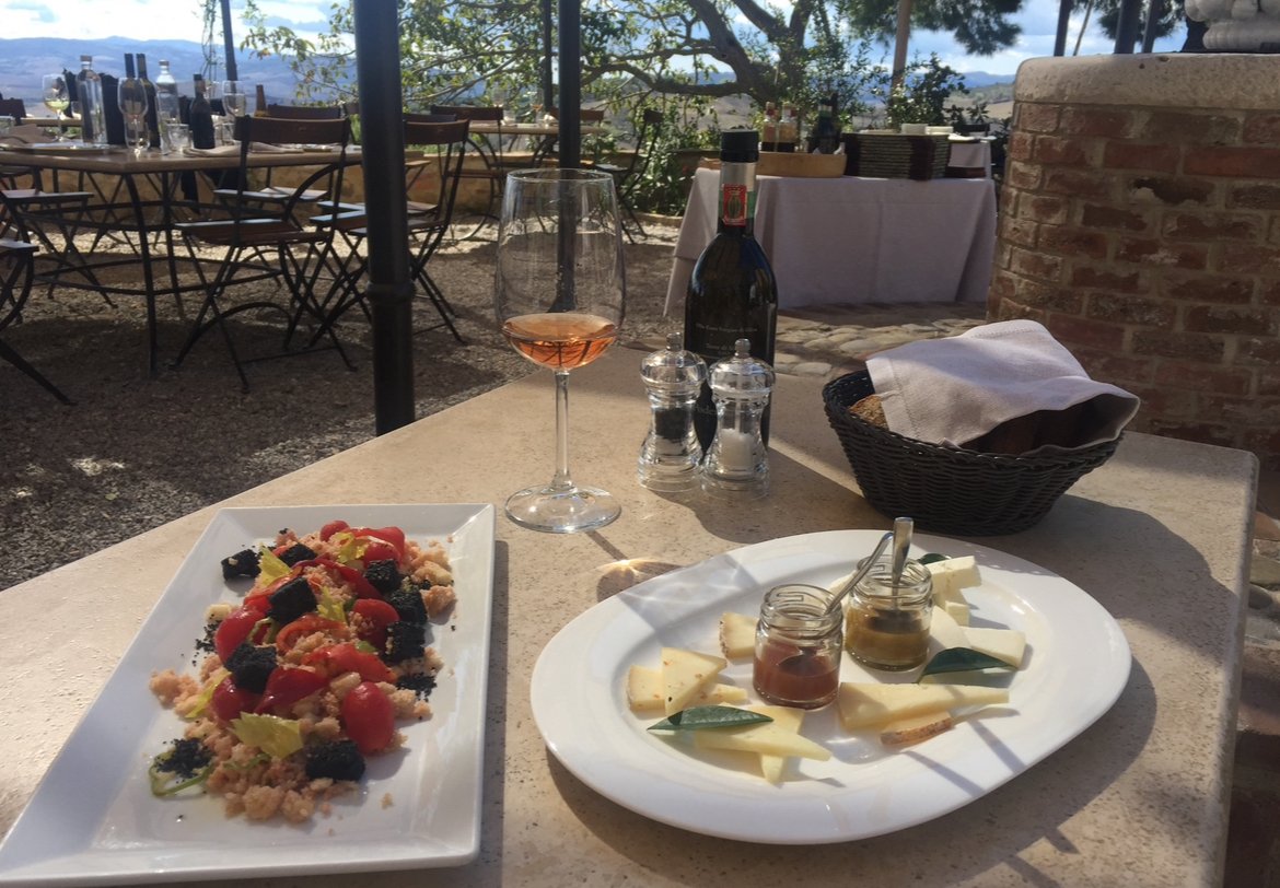 Table with a glass of rosé wine, a bottle of red wine, salt and pepper shakers, a basket of napkins, a white plate with cheese, green leaves, and small jars of honey and jam, and a rectangular dish with a salad containing cherry tomatoes, blackberries, and lettuce, at an outdoor restaurant with tables and chairs and trees in the background.