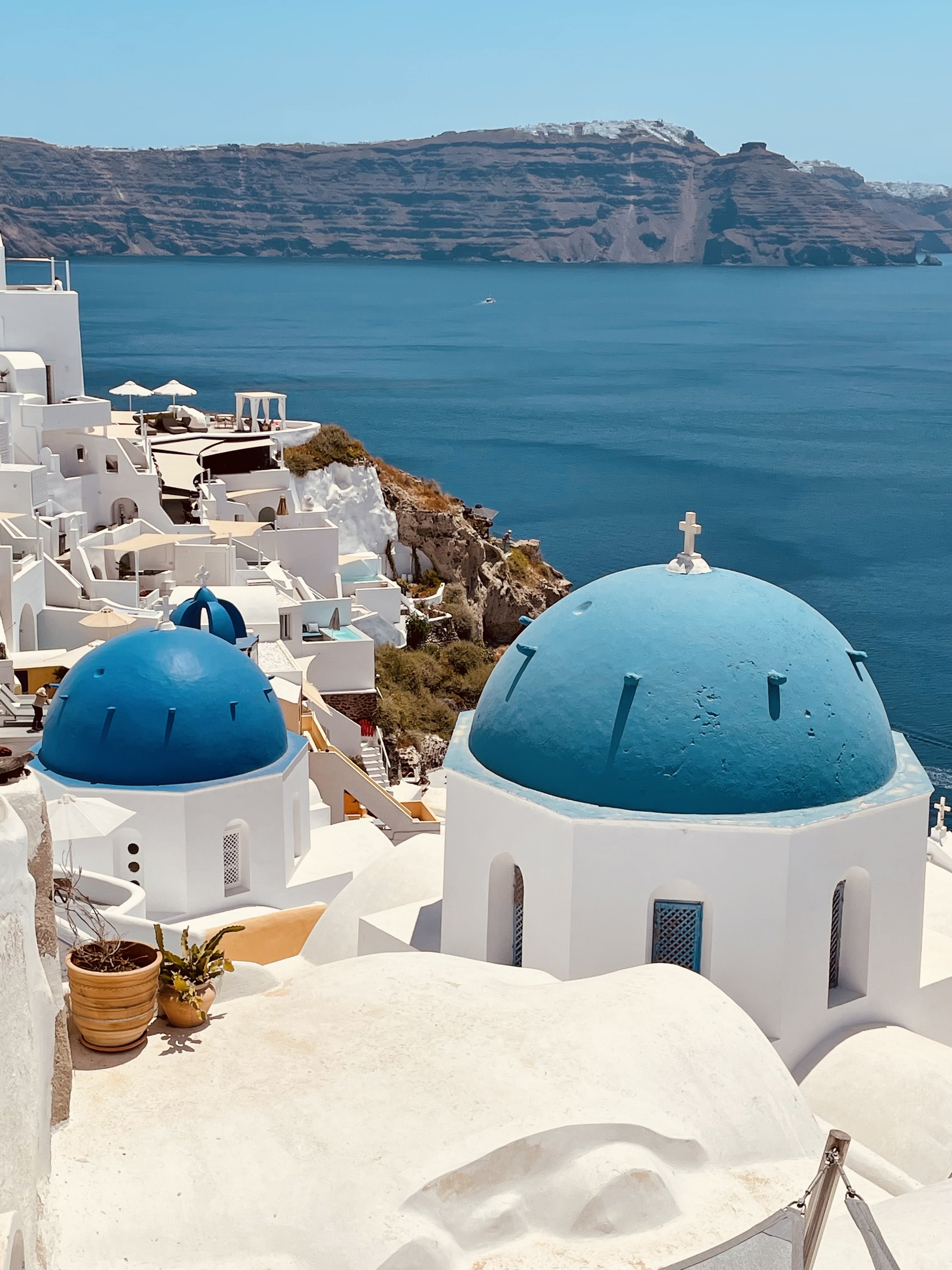 White buildings with blue domes on a cliffside overlooking the sea in Santorini, Greece, with a distant landform across the water.