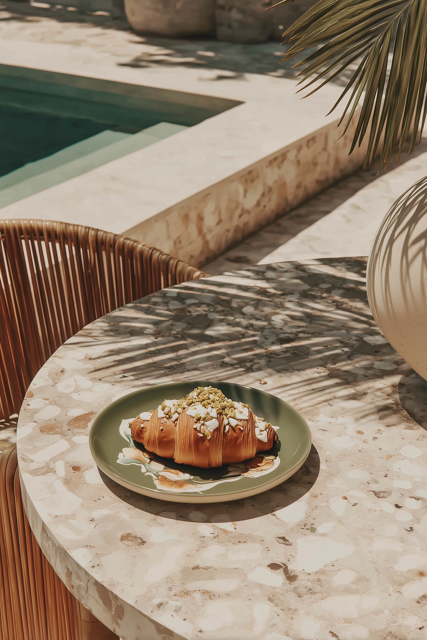 A croissant pastry topped with white icing, crushed pistachios, and crumbled white chocolate on a green ceramic plate, placed on a marble outdoor table near a pool.