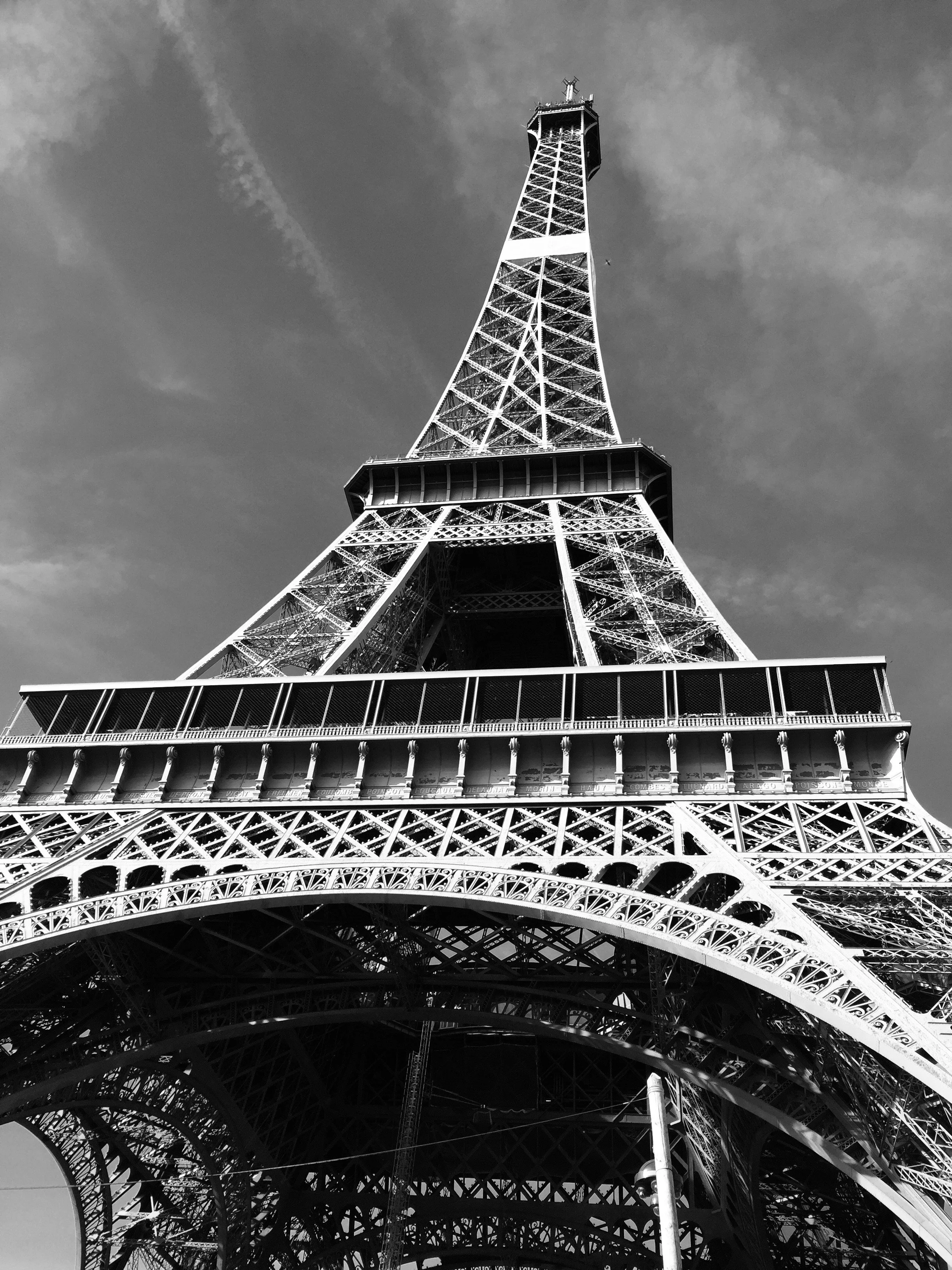 Black and white photo of the Eiffel Tower from below, highlighting its intricate iron lattice structure against a partly cloudy sky.