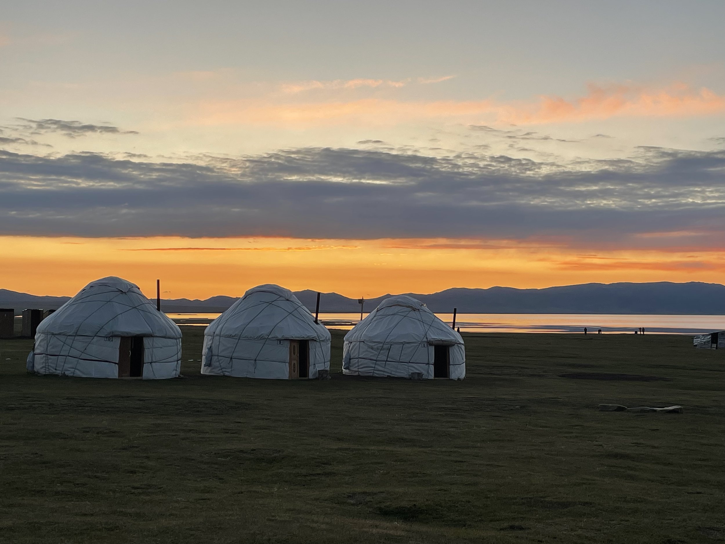 Three traditional yurts on a grassy field at sunset, overlooking a body of water and mountains in the distance.