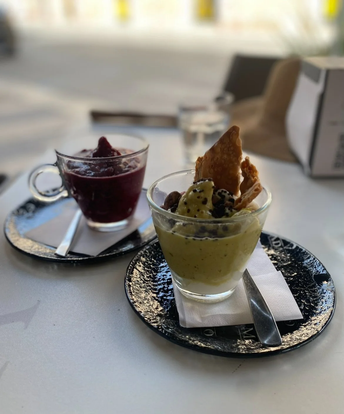 Dessert cups with ice cream and fruit on decorative plates, with a spoon and napkin underneath, on a white table.