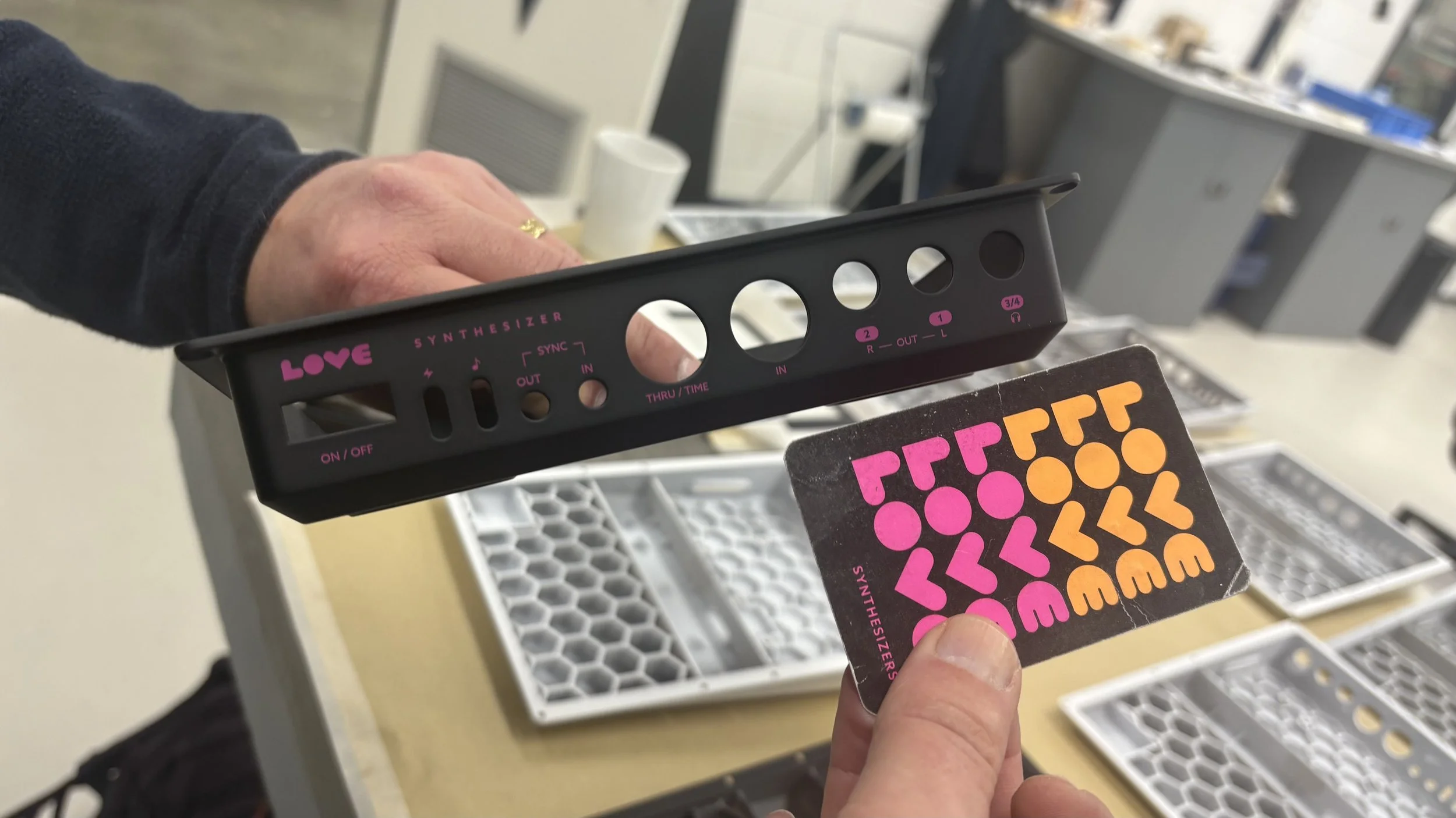 A person's hand holding a black metal object with pink lettering and various holes, next to a card with colorful geometric shapes, in a room with display shelves and trays containing similar metal objects.