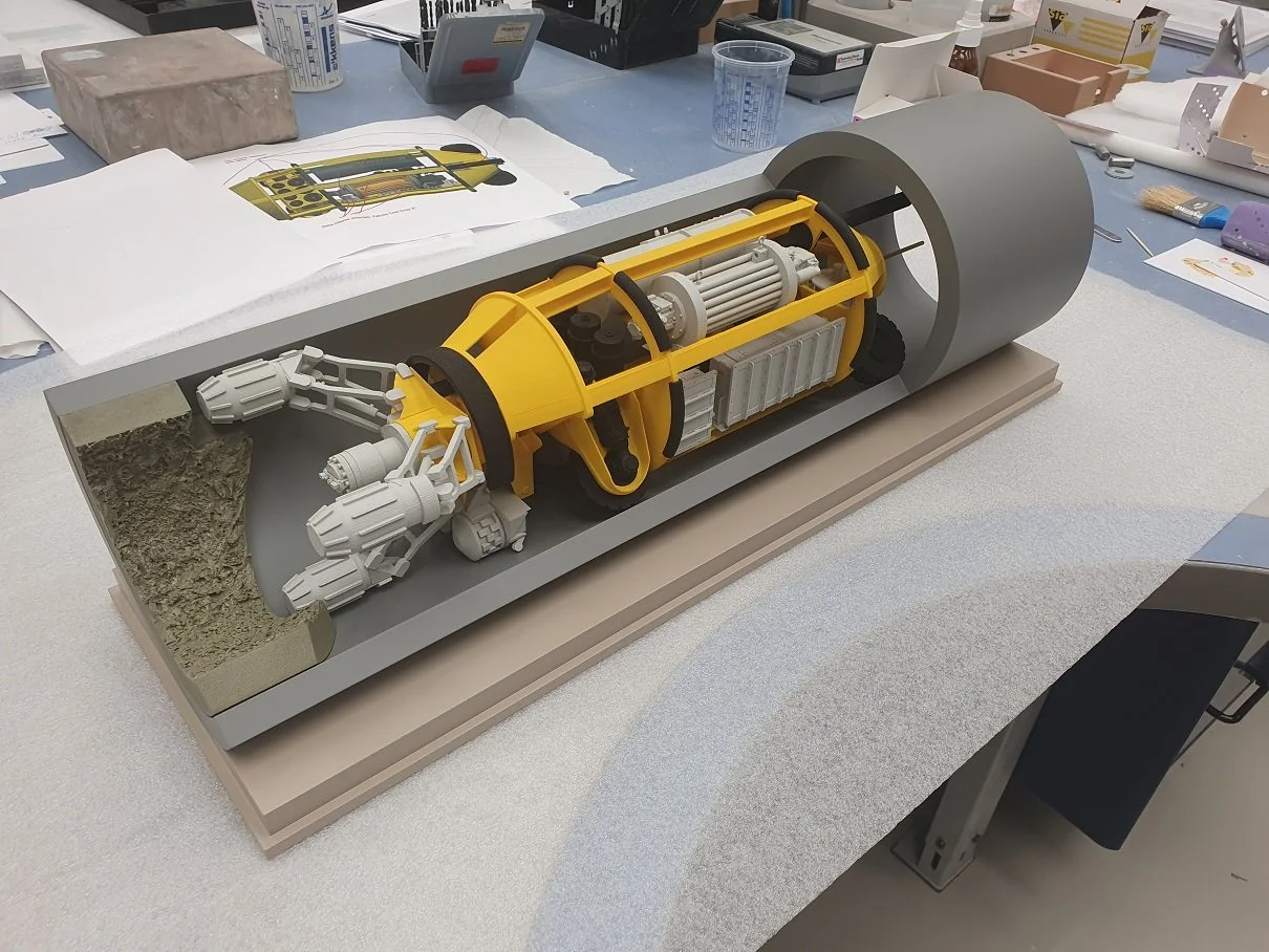 A detailed cross-section model of a machine or engine with yellow housing, white mechanical parts, and a large gray cylindrical section, displayed on a workbench with papers and tools around.