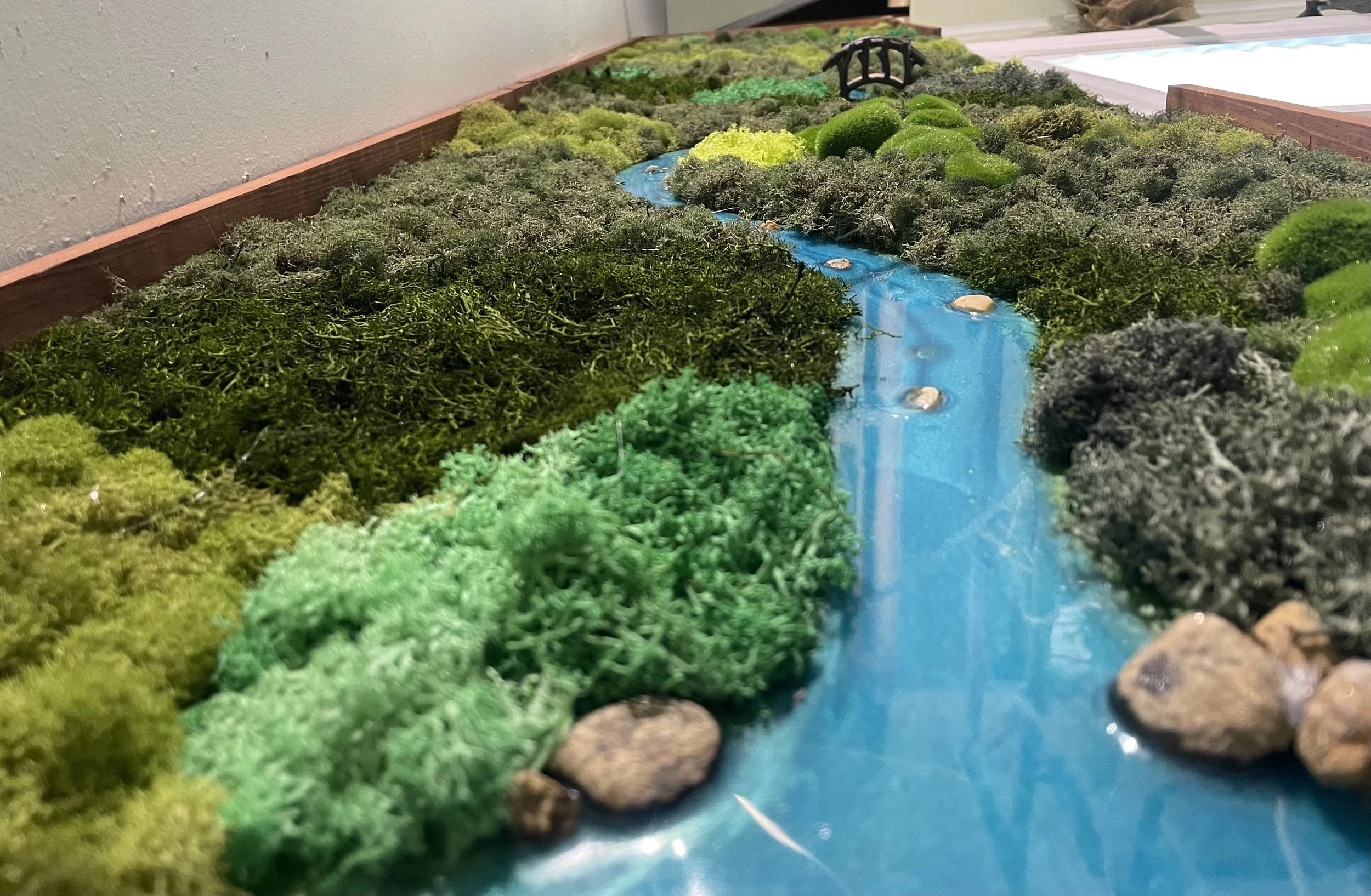 Miniature landscape featuring a winding blue river made of shiny material, surrounded by green moss, artificial grass, and small rocks, with a tiny black bridge structure in the background.