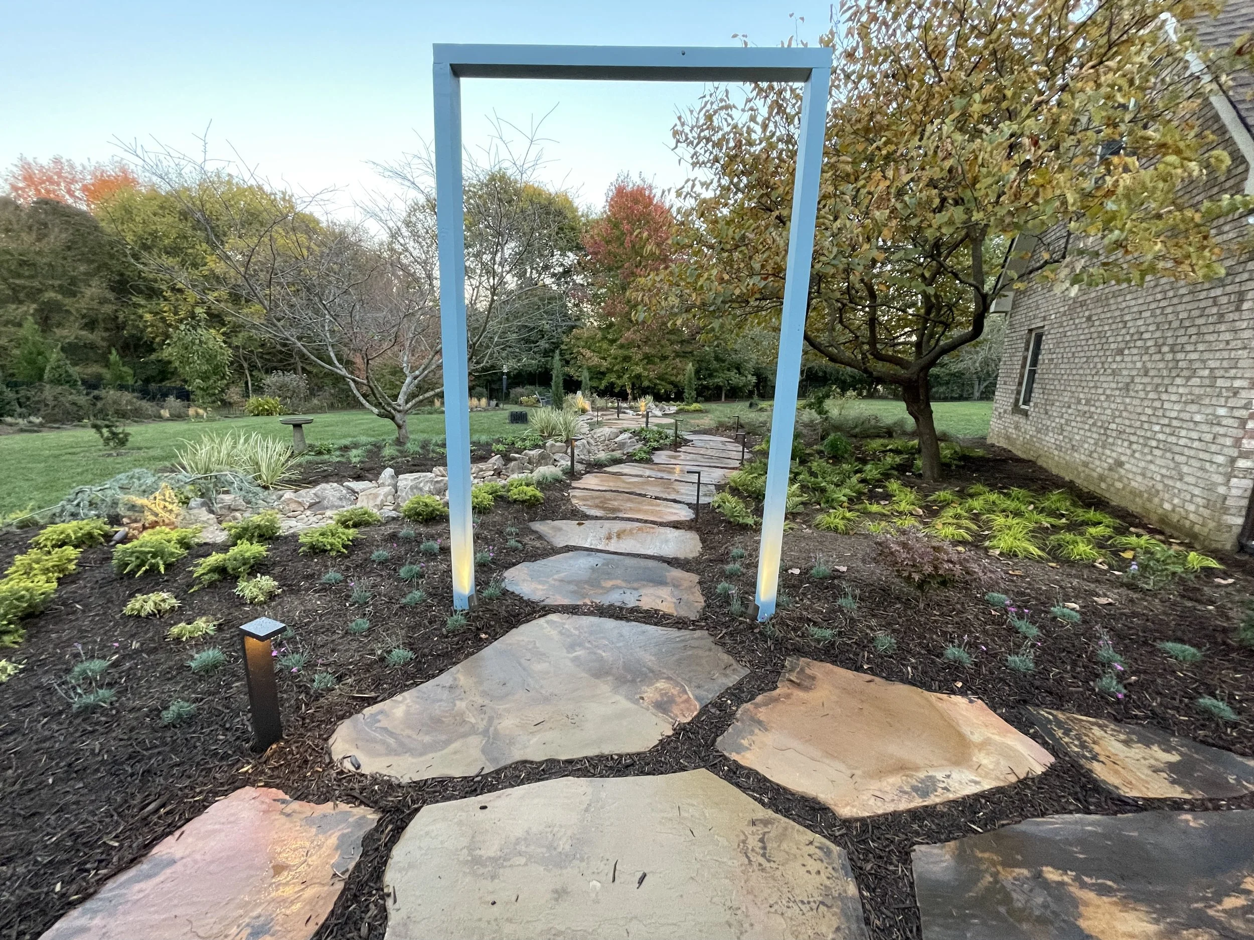 Stone pathway leading through a landscaped garden with small plants, trees, and a brick building on the right, under a light blue frame structure.