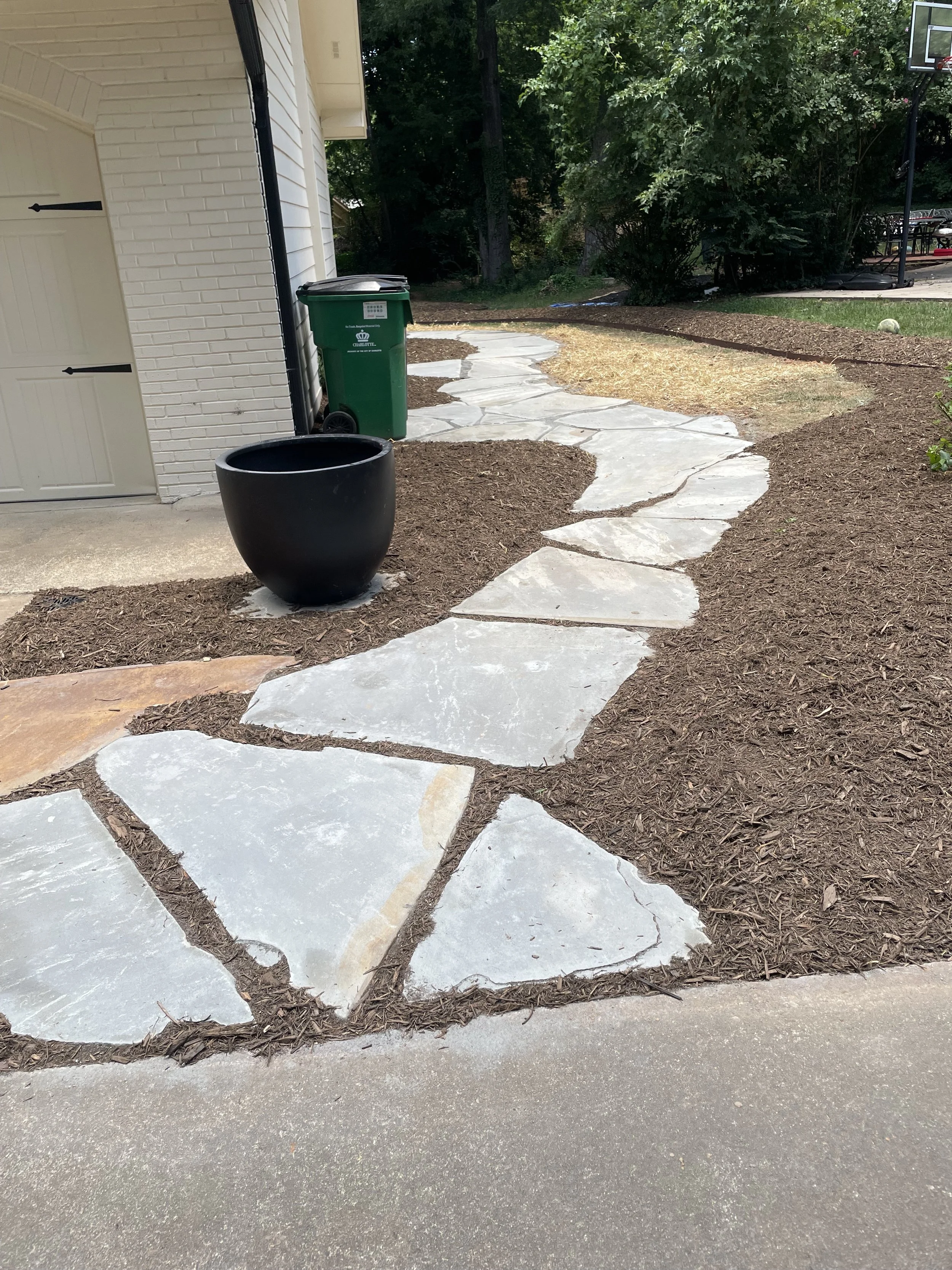 Stone pathway in a yard with mulch on either side, leading away from a house with a brick wall, a green trash bin, and a large black planter
