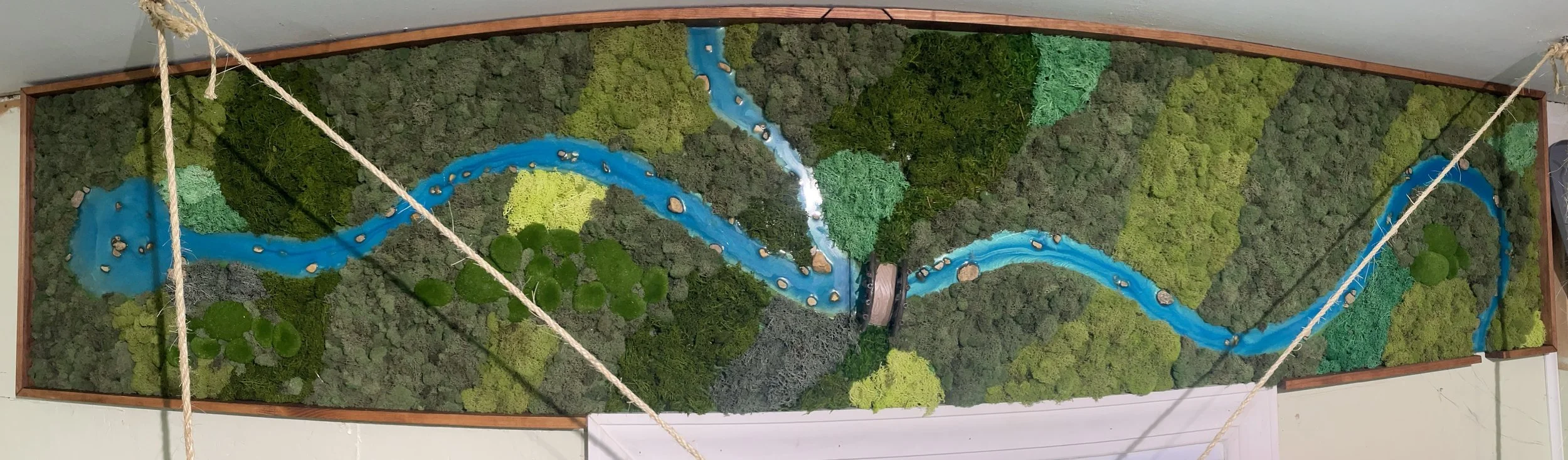 A vertical wall mural depicting a lush forest landscape with a blue river flowing through it, surrounded by various green moss and plant textures.