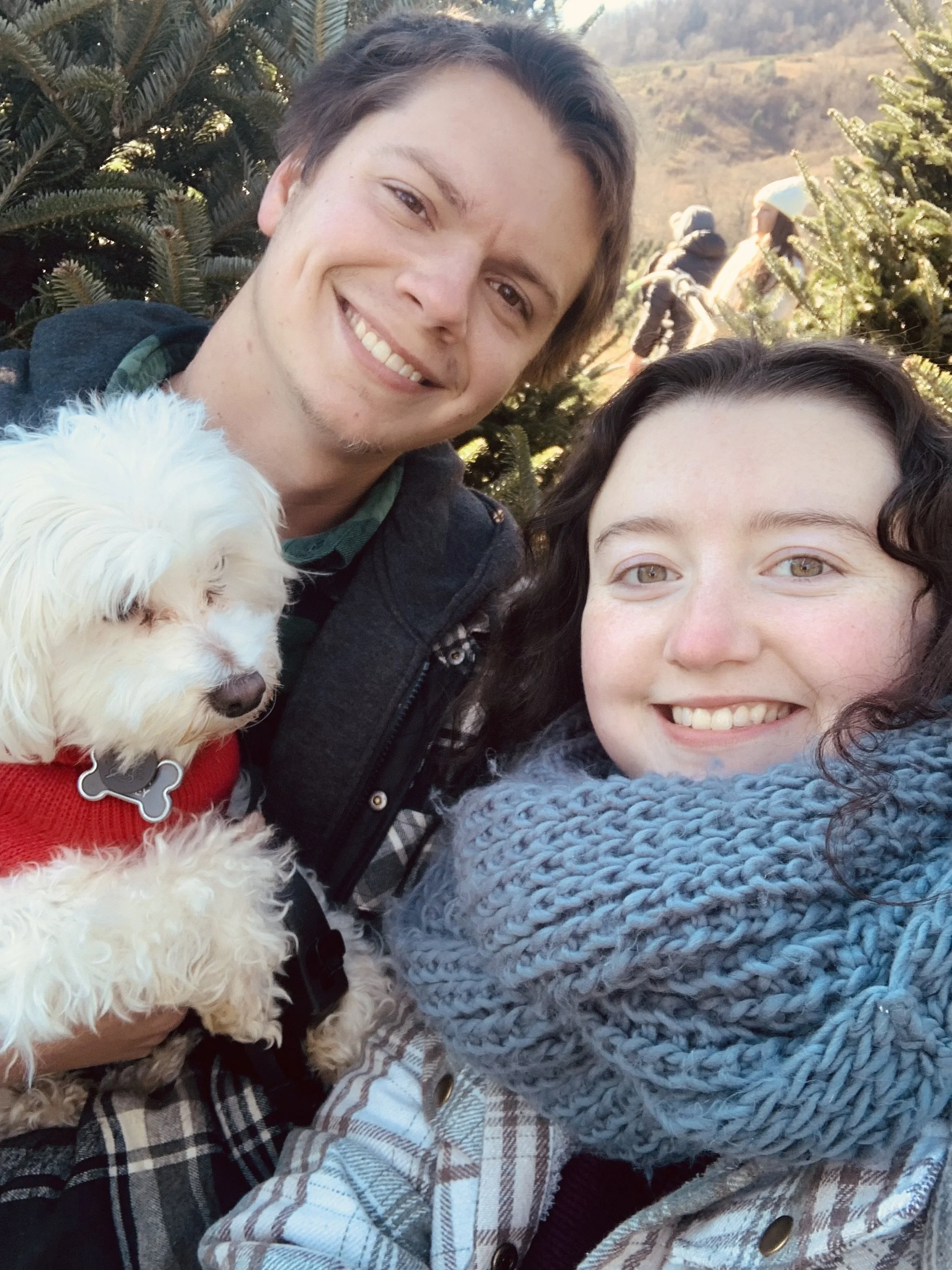 A smiling young couple taking a selfie outdoors during daytime, with a small white dog wearing a red sweater in the man's arm, standing in front of green pine trees with a background of distant hills and a few blurred people.