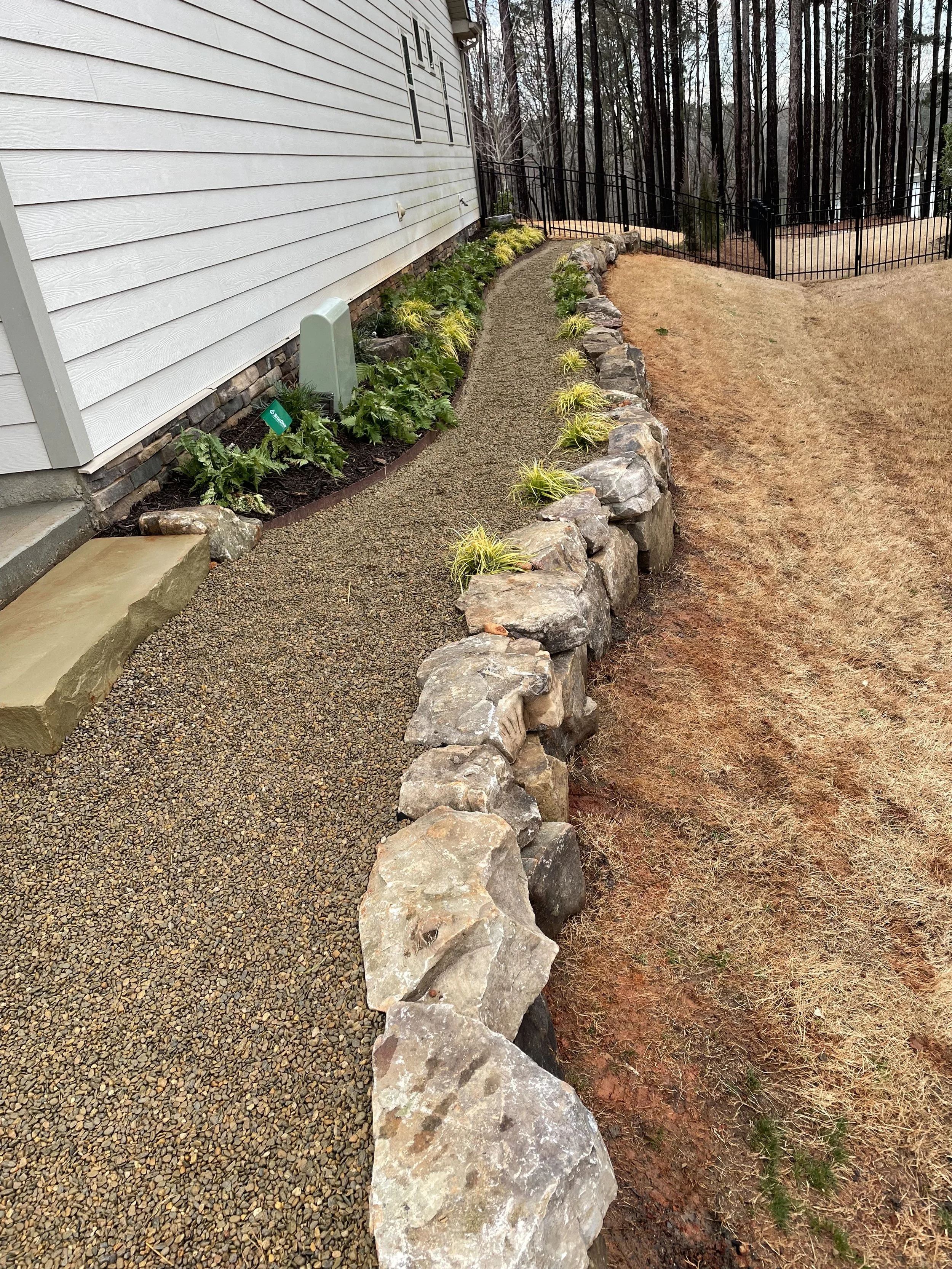 A side yard with a gravel pathway bordered by large rocks, a white house with horizontal siding, and a garden bed with plants. In the background, there is a black metal fence and a wooded area.