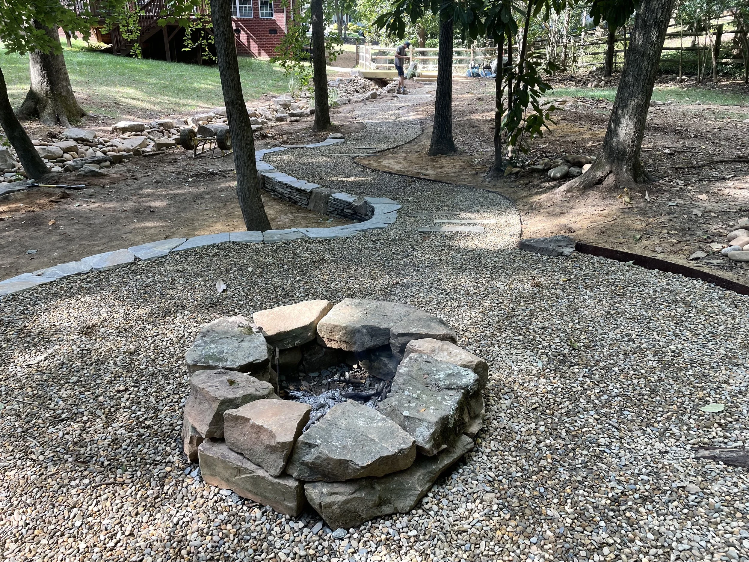 A backyard with a small fire pit made of large stones, a gravel pathway winding through trees, and a person in the background near a raised deck.