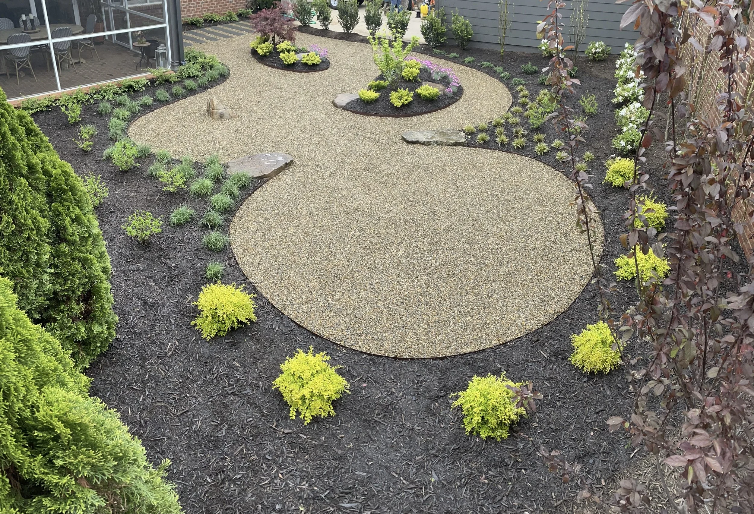 Garden with a winding concrete pathway, surrounded by various plants, shrubs, small trees, and mulch. There is a patio with chairs and a glass-enclosed room in the background.