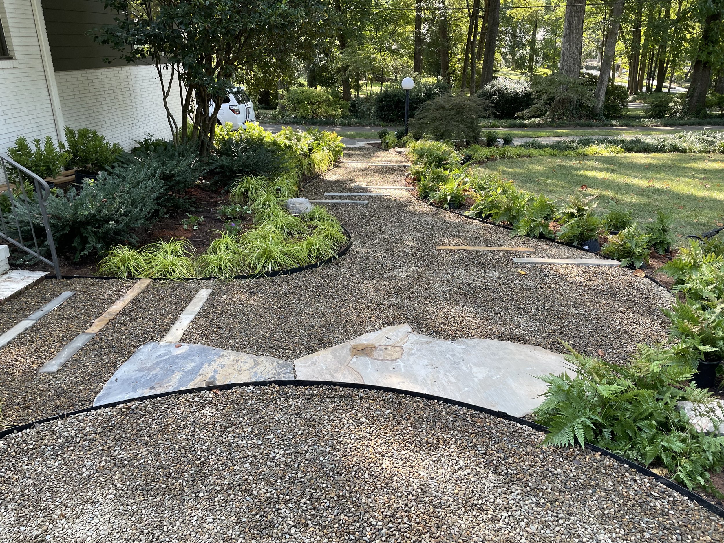 A gravel pathway bordered by lush green bushes and plants, leading to a garden with a lamp post, surrounded by trees and greenery in a residential yard.