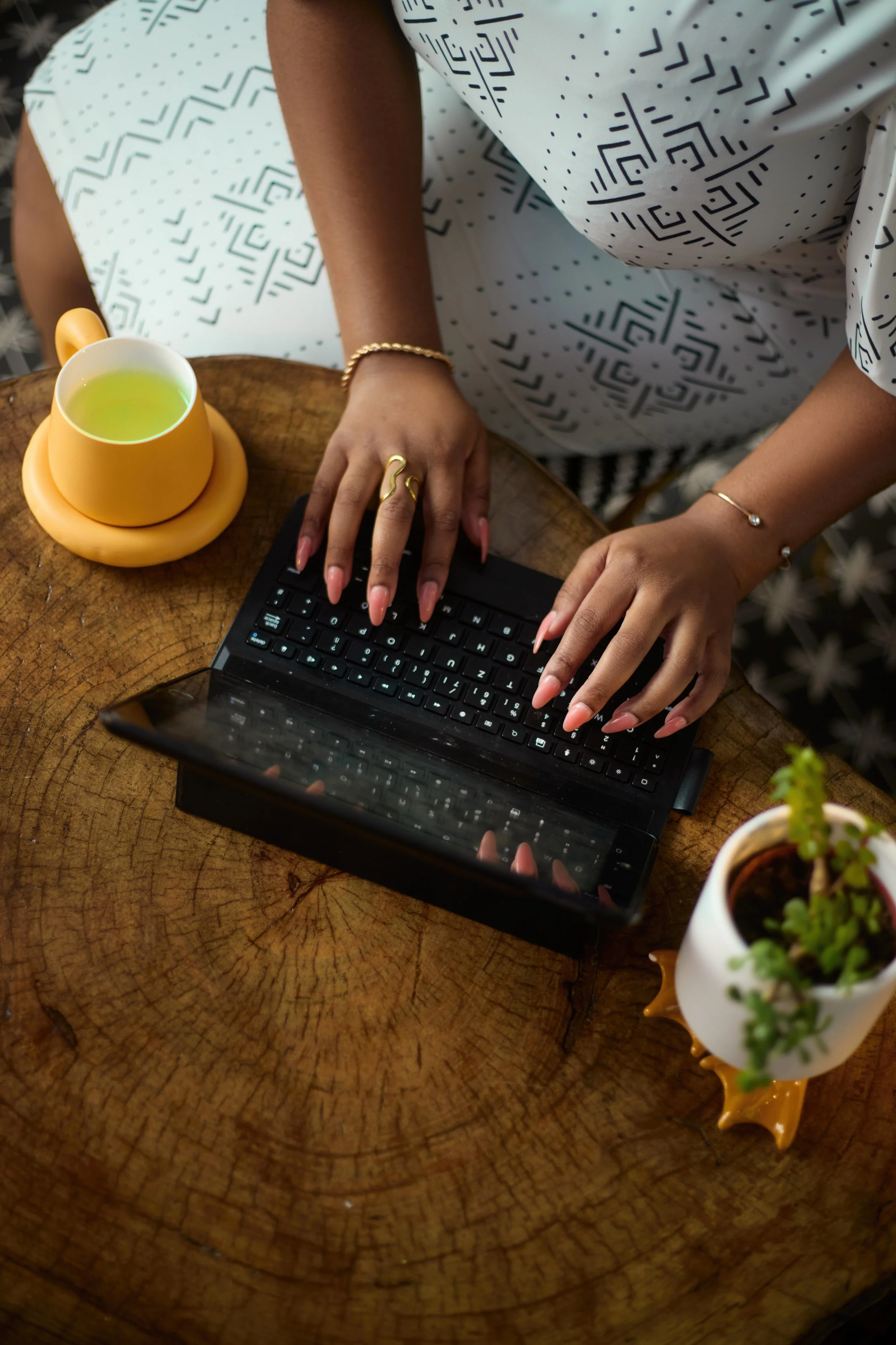 Une personne tapant sur un clavier d'ordinateur portable posé sur une table en bois avec une tasse de thé vert, une petite plante et un vase de fleurs.