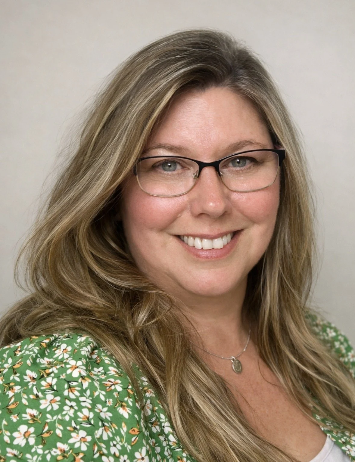 A woman with long blonde hair and glasses, smiling at the camera, wearing a green floral top and a necklace.
