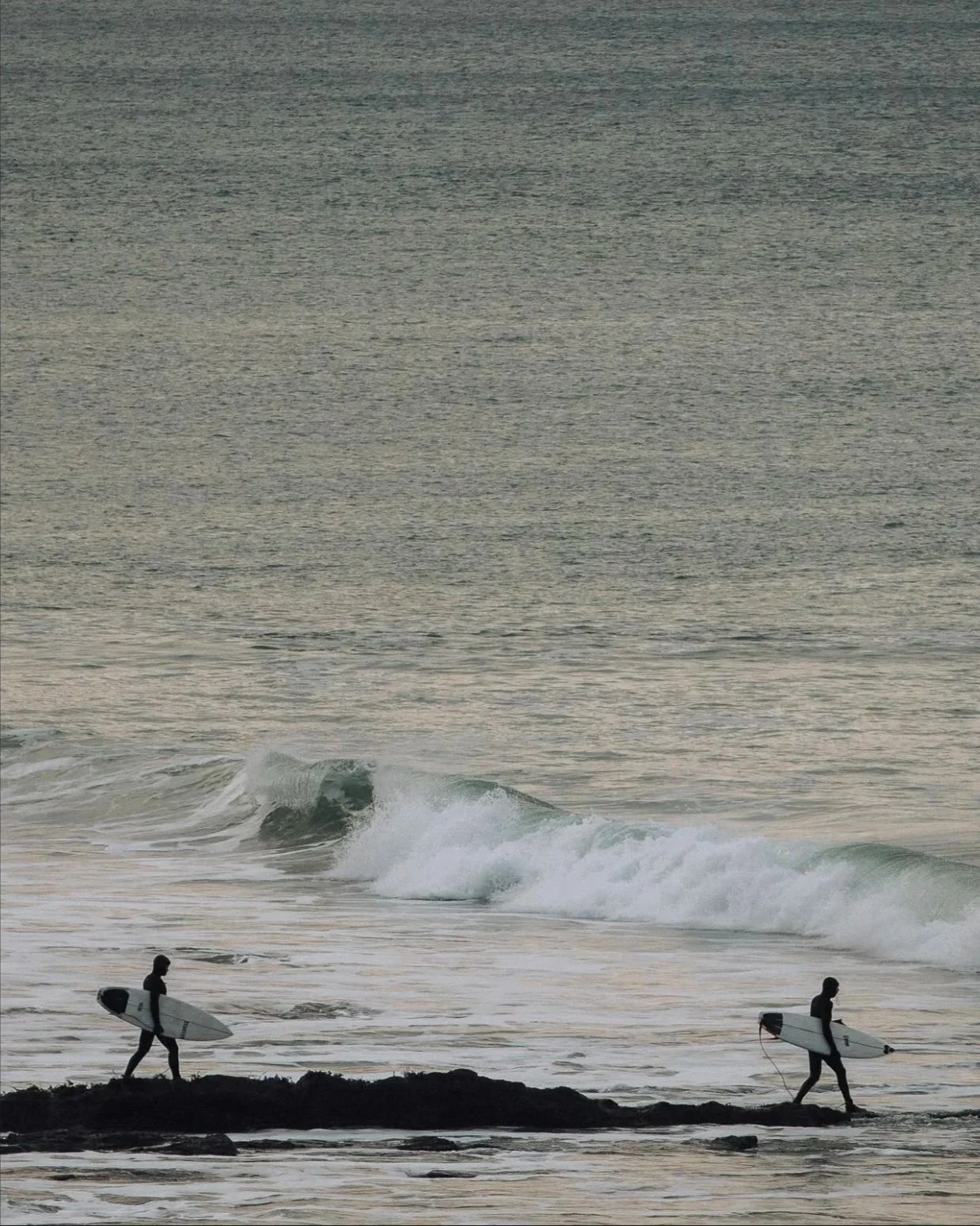Two surfers walking on rocks toward the ocean, each carrying a surfboard.