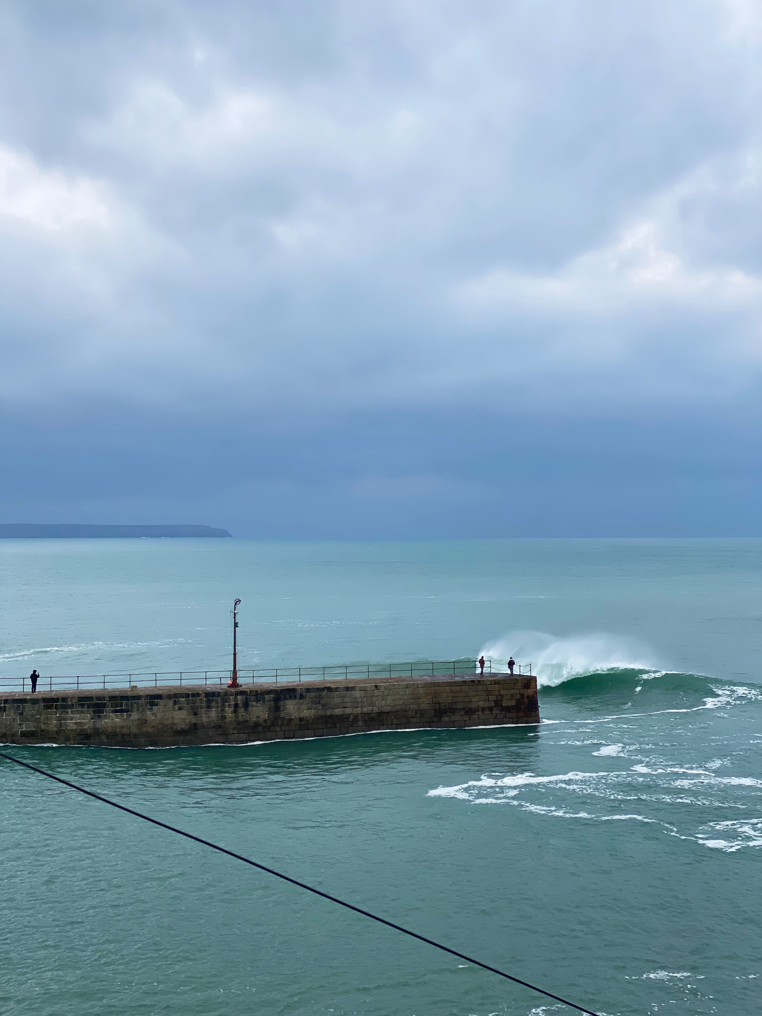 A stone pier extending into the ocean with a few people standing at the edge, and waves crashing against the pier under a cloudy sky.