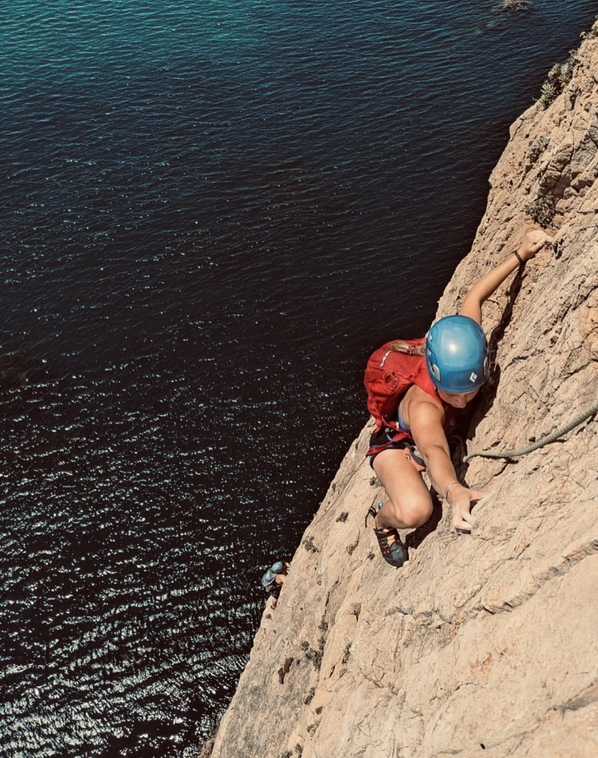 Person wearing a blue helmet and red backpack rock climbing on a steep cliff near water.