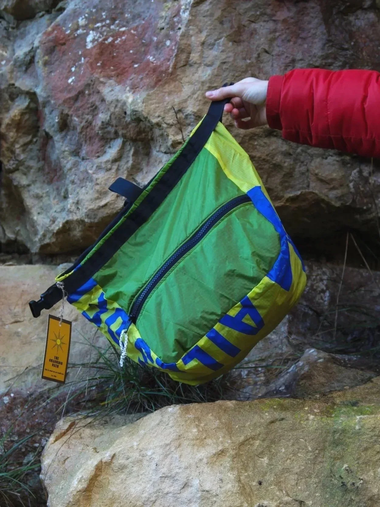 A person wearing a red jacket holds a green, blue, and yellow chalk bag with a zipper in front of a rocky background.