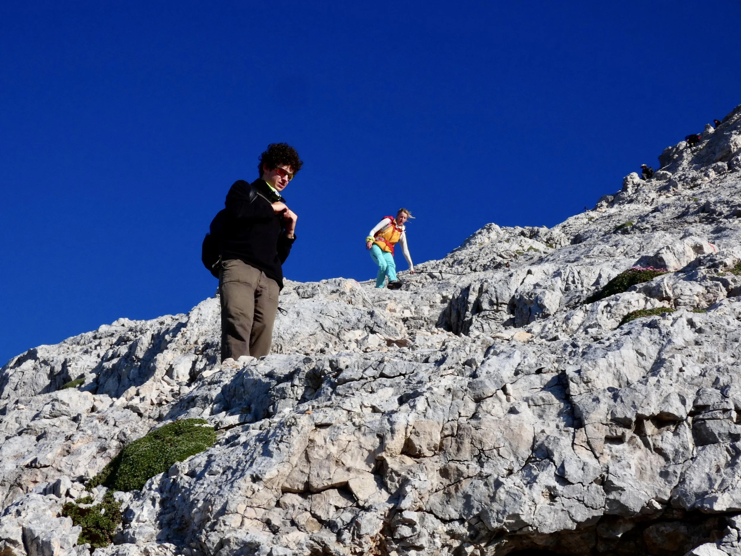 Two hikers trekking on rocky mountain terrain under a clear blue sky.